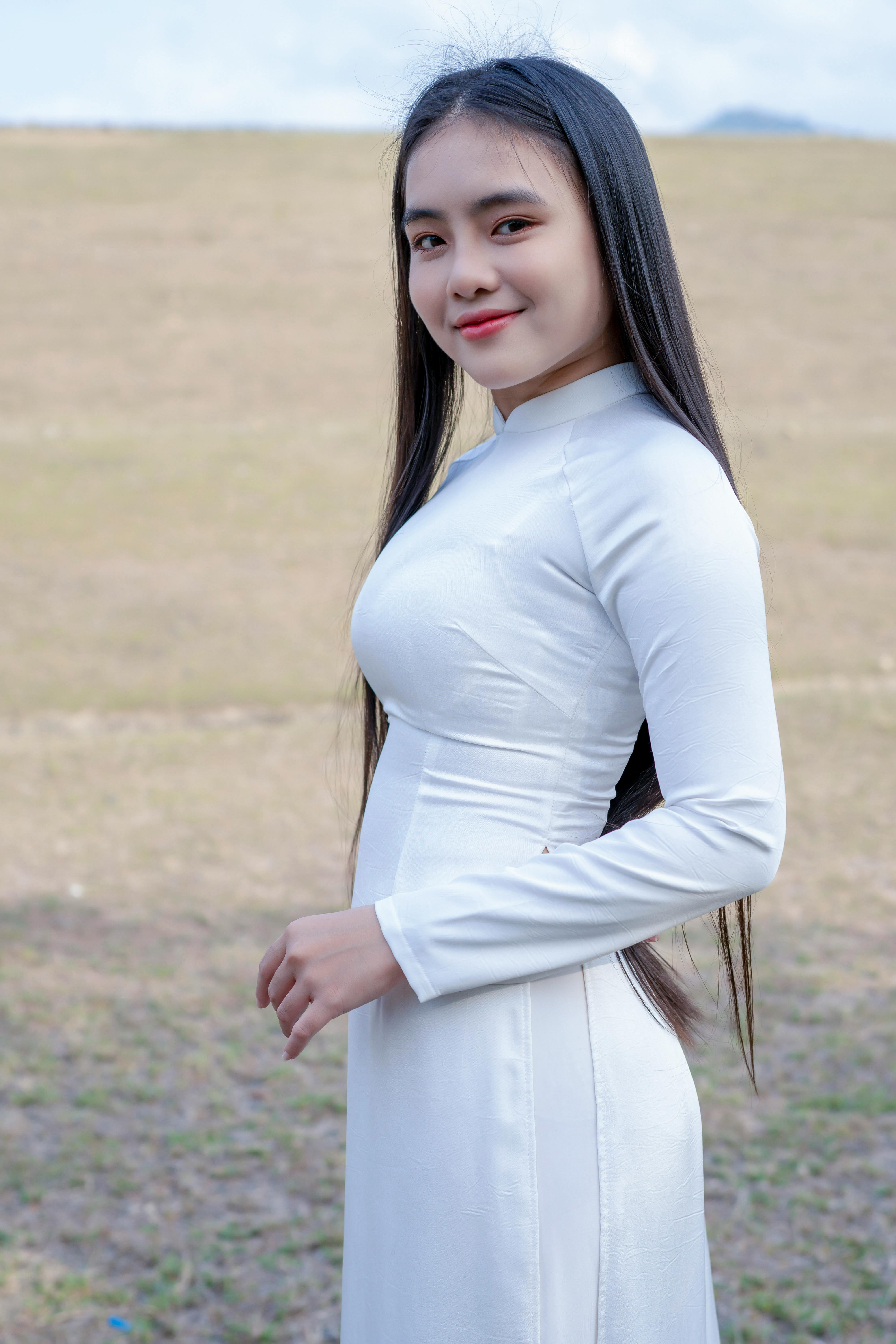 Woman in white dress standing in a field, looking towards the camera with a smile.