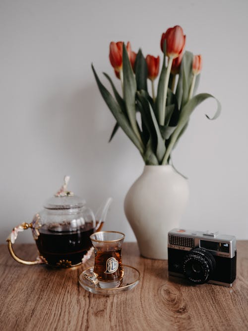 A Glass Pot with Tea and a Glass of Tea Standing on a Table with Flowers and a Camera 