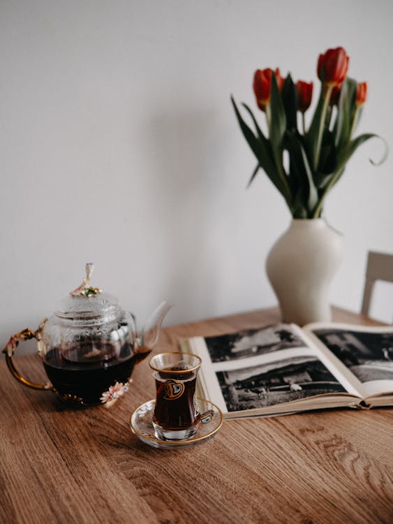 A Glass Pot with Tea and a Glass of Tea Standing on a Table with Flowers and a Magazine 