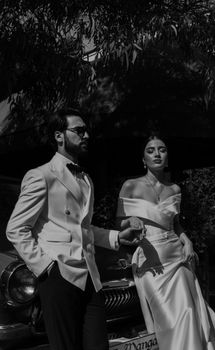 Elegantly dressed couple in black and white wedding photography, standing by a vintage car.