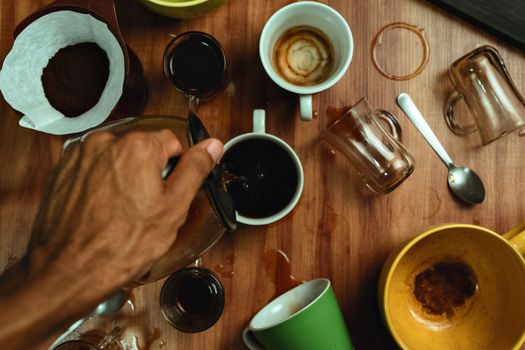 Top view of a hand pouring coffee into a mug surrounded by spilled cups on a wooden table.