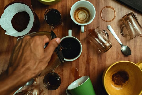 Top view of a hand pouring coffee into a mug surrounded by spilled cups on a wooden table.