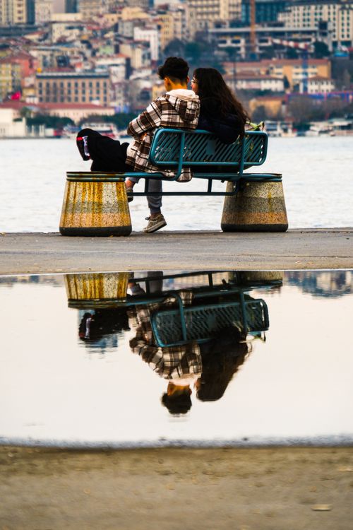 Free A couple sits on a bench by the city shore, reflecting urban life in a puddle. Stock Photo