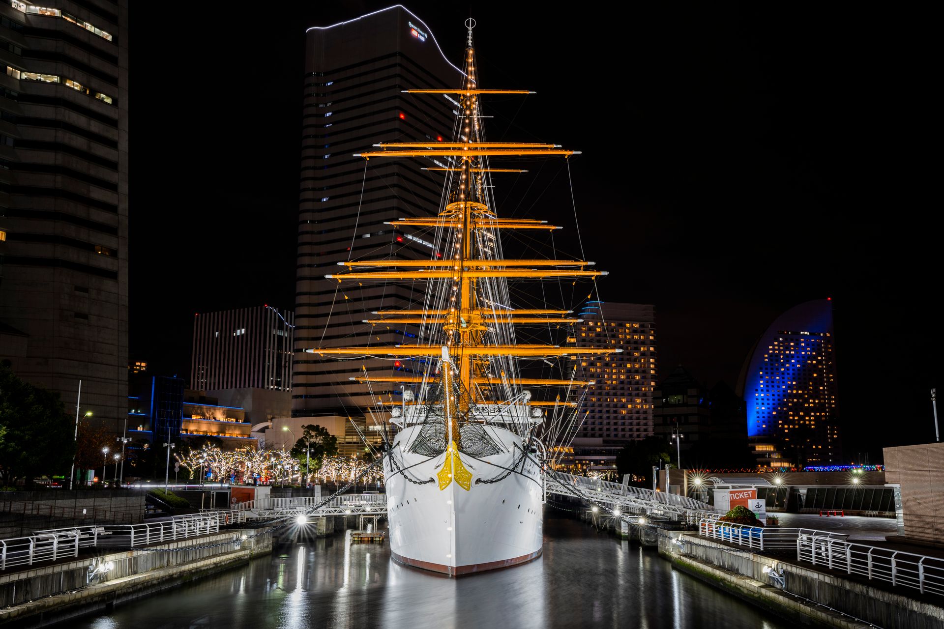 Illuminated Nippon Maru ship docked in Yokohama harbor surrounded by urban skyline at night.