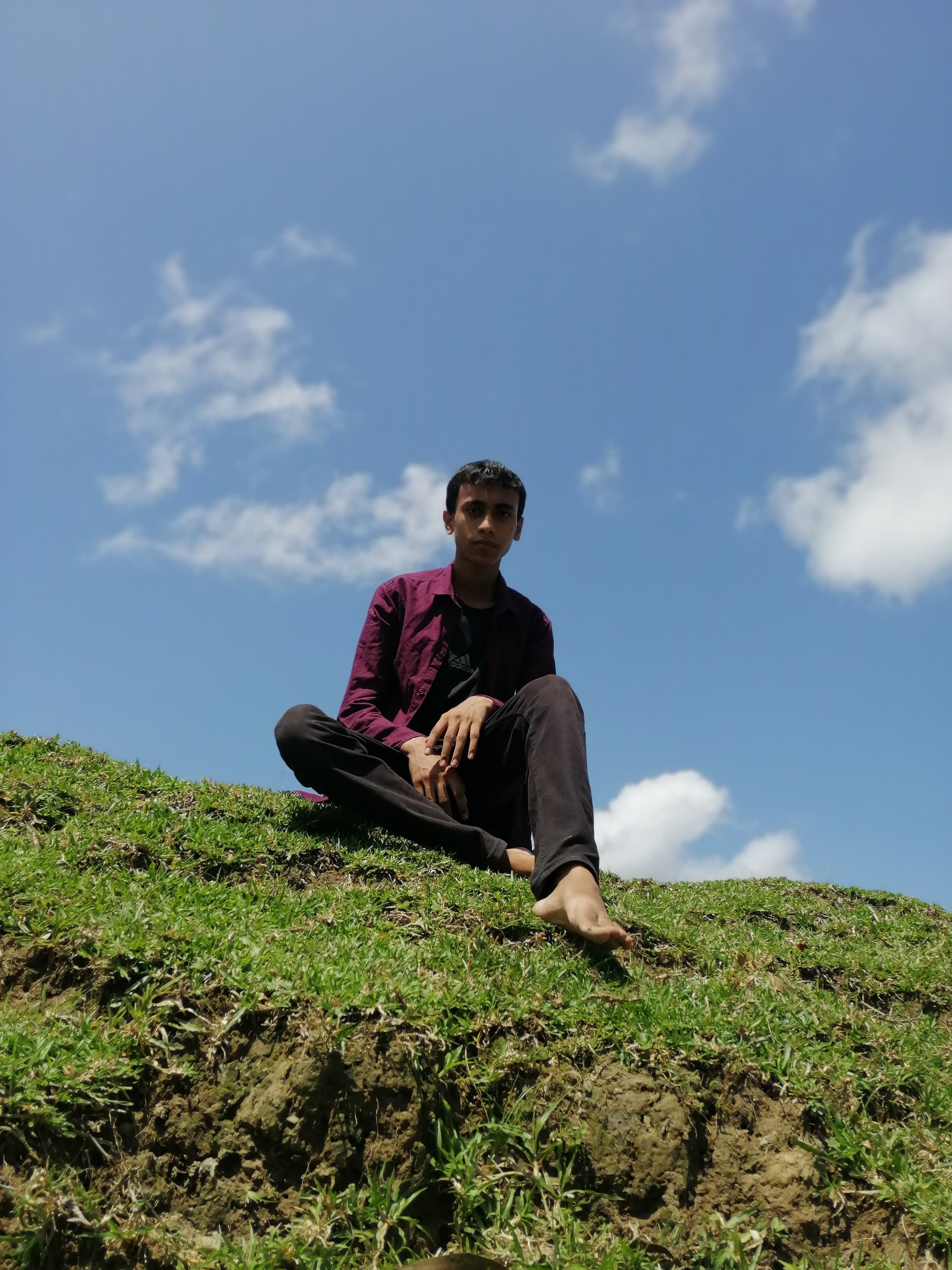 Barefoot young man sitting on grass under blue sky, enjoying summer outdoors.