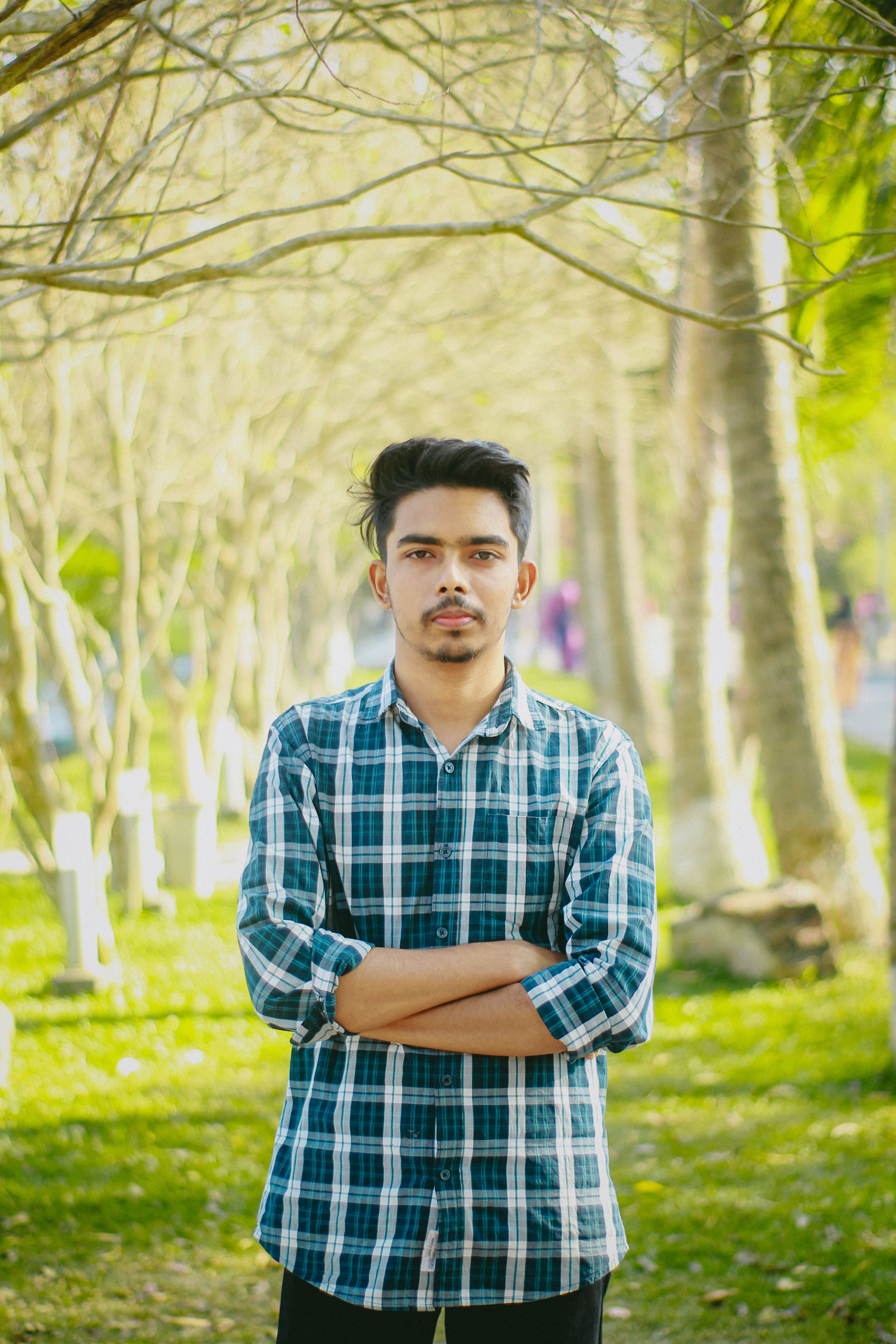 A young man stands confidently in a park with arms crossed, surrounded by trees.