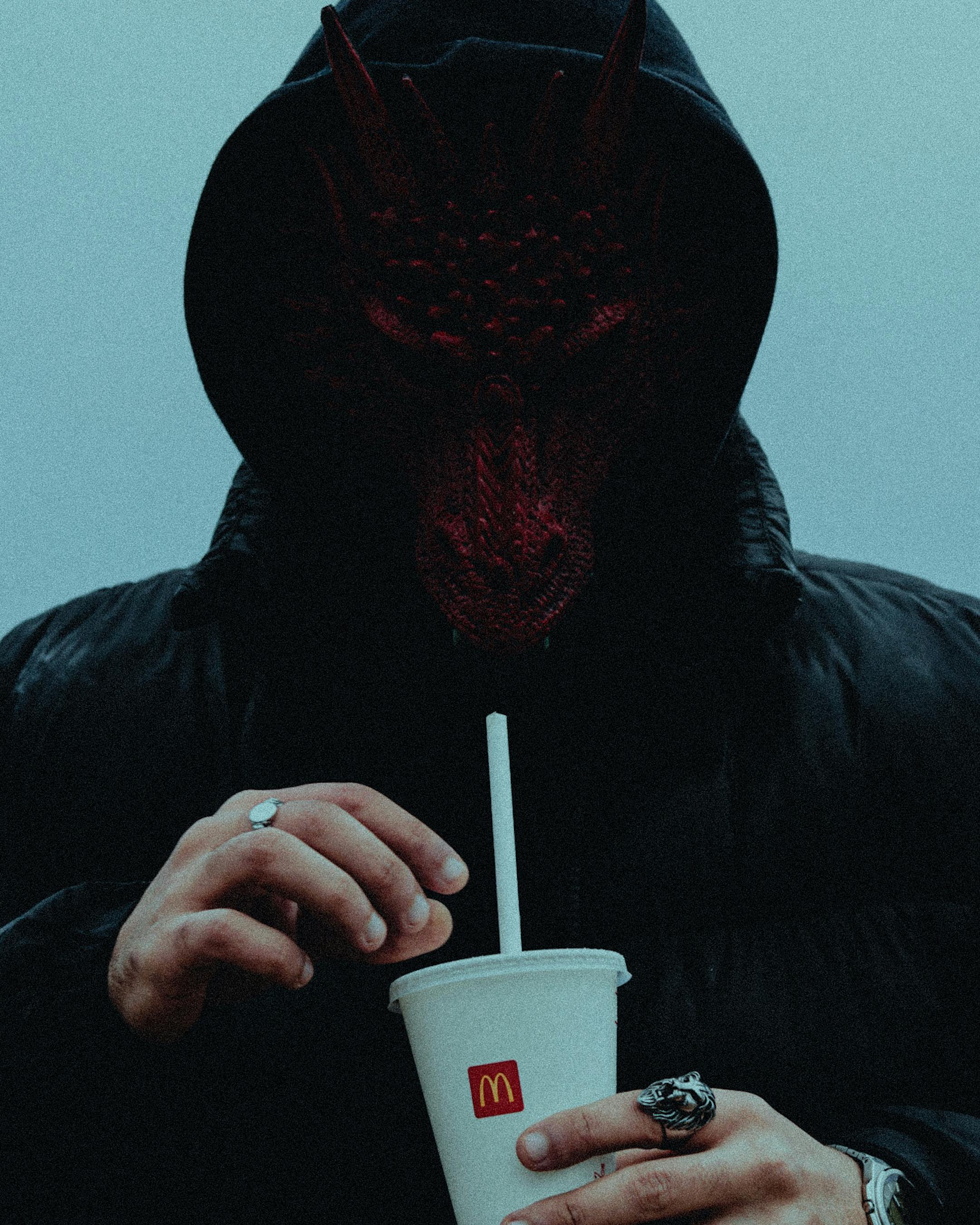 A person in a dark mask holding a McDonald's cup outdoors. Eerie atmosphere.