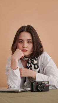 Stylish woman in a thoughtful pose with a retro camera and laptop on a warm colored background.