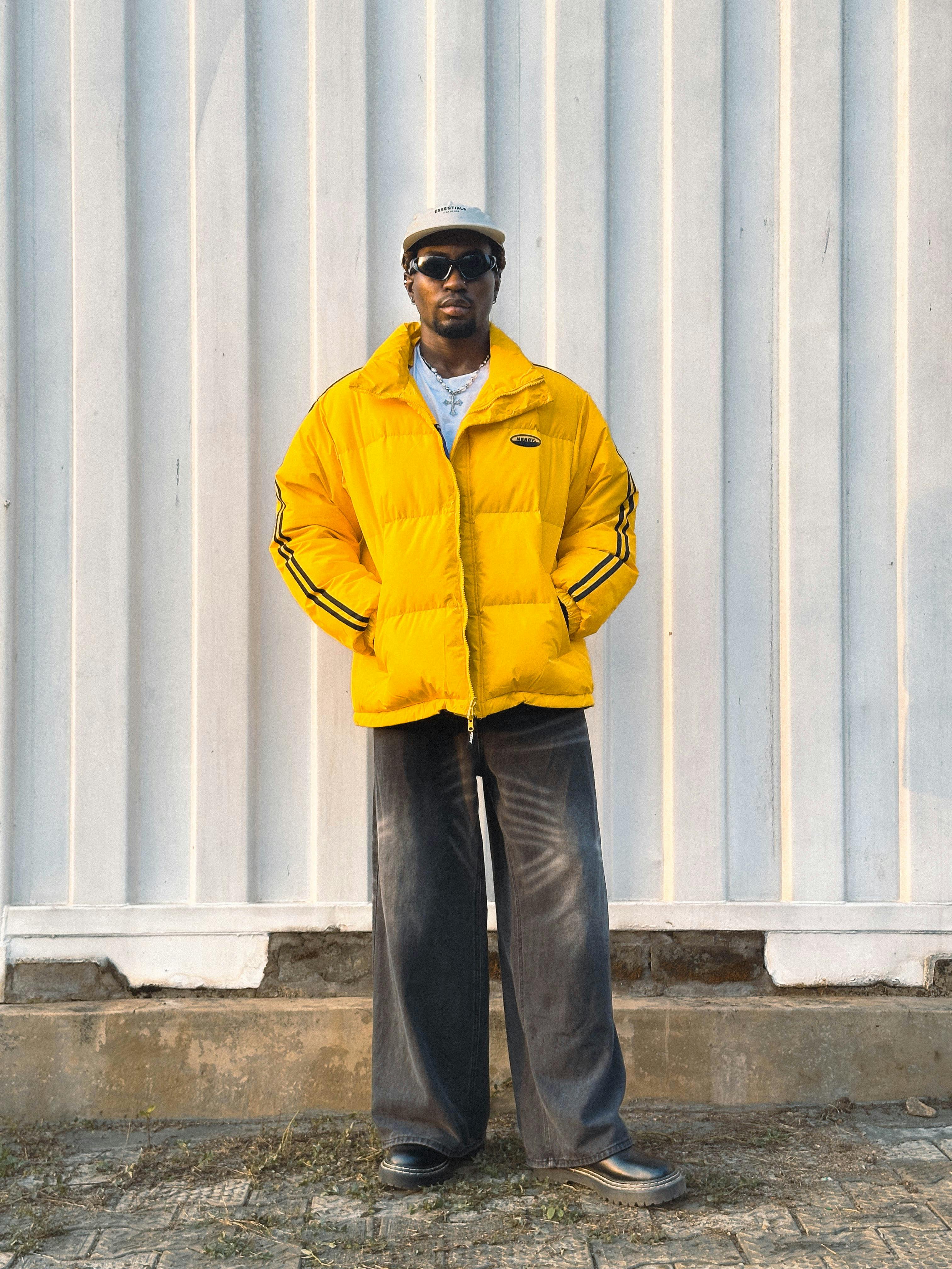 Man wearing a yellow puffer jacket and sunglasses, posing outdoors against a white wall.