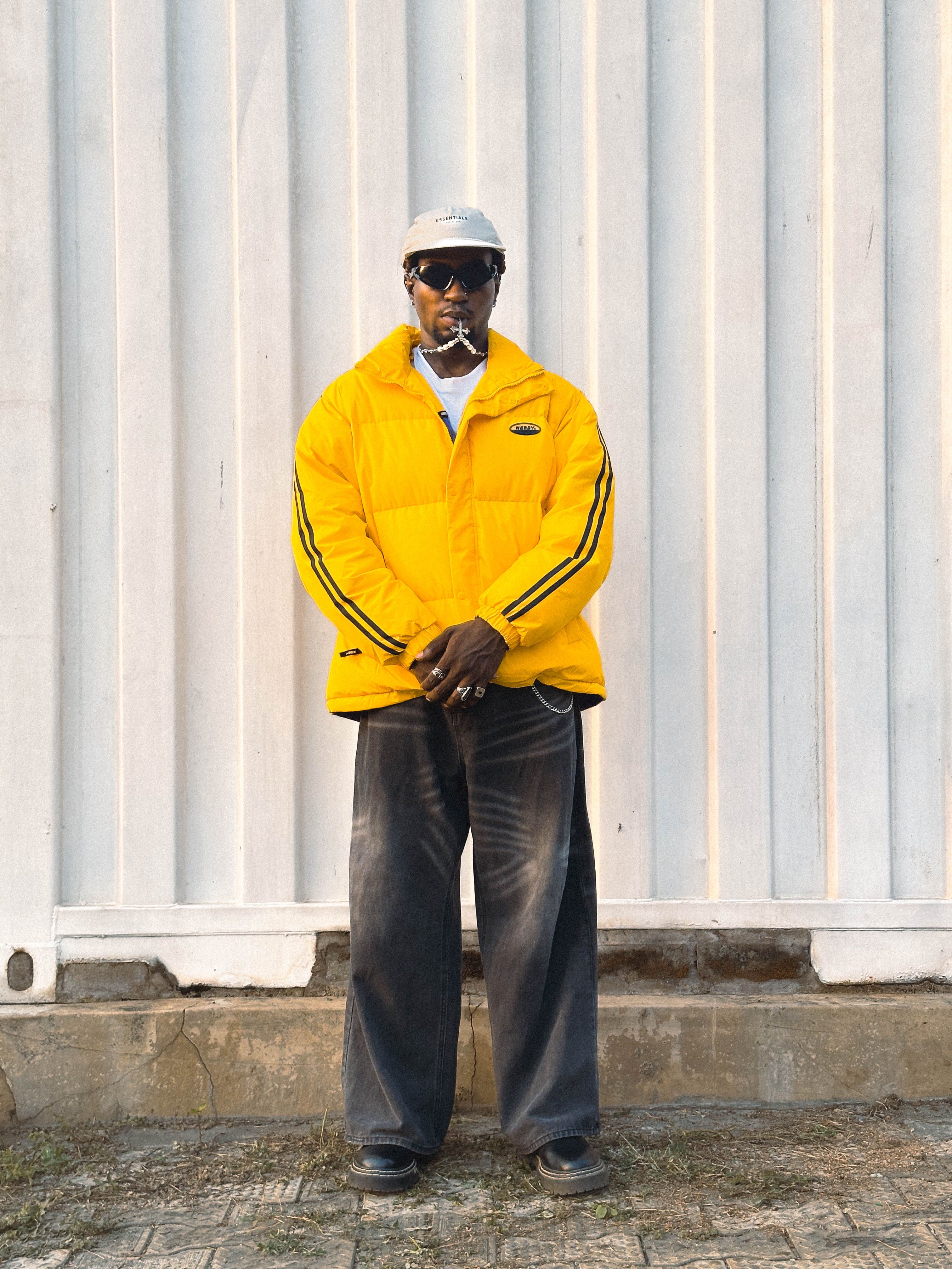 A fashionable man in a yellow jacket poses confidently against a textured wall, showcasing urban style.