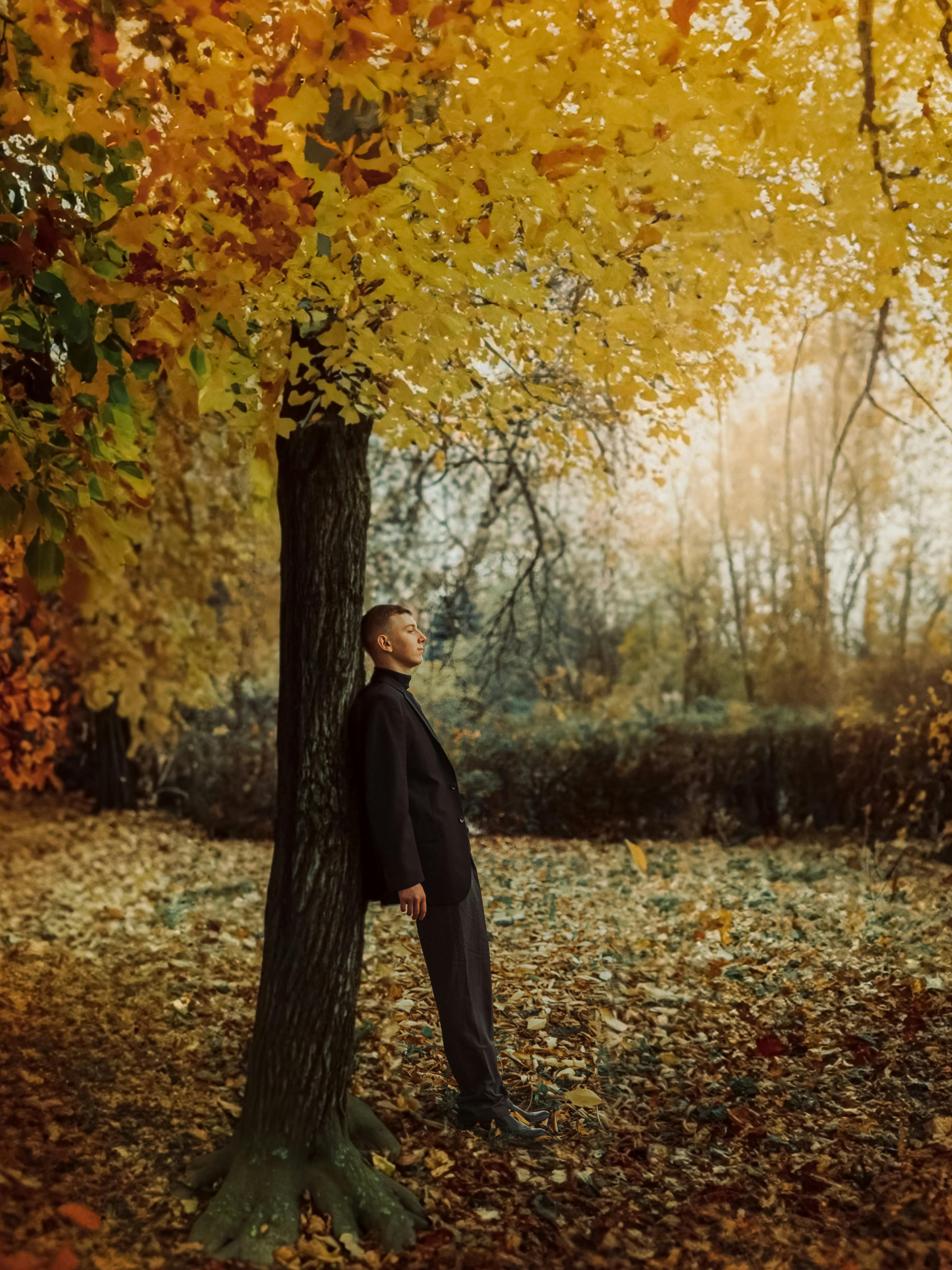 Young man in a suit leaning against a tree in a vibrant autumn park.
