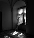 这是一张黑白照片，一名身着连衣裙的女子站在窗前，在强烈的光影对比下拍摄而成