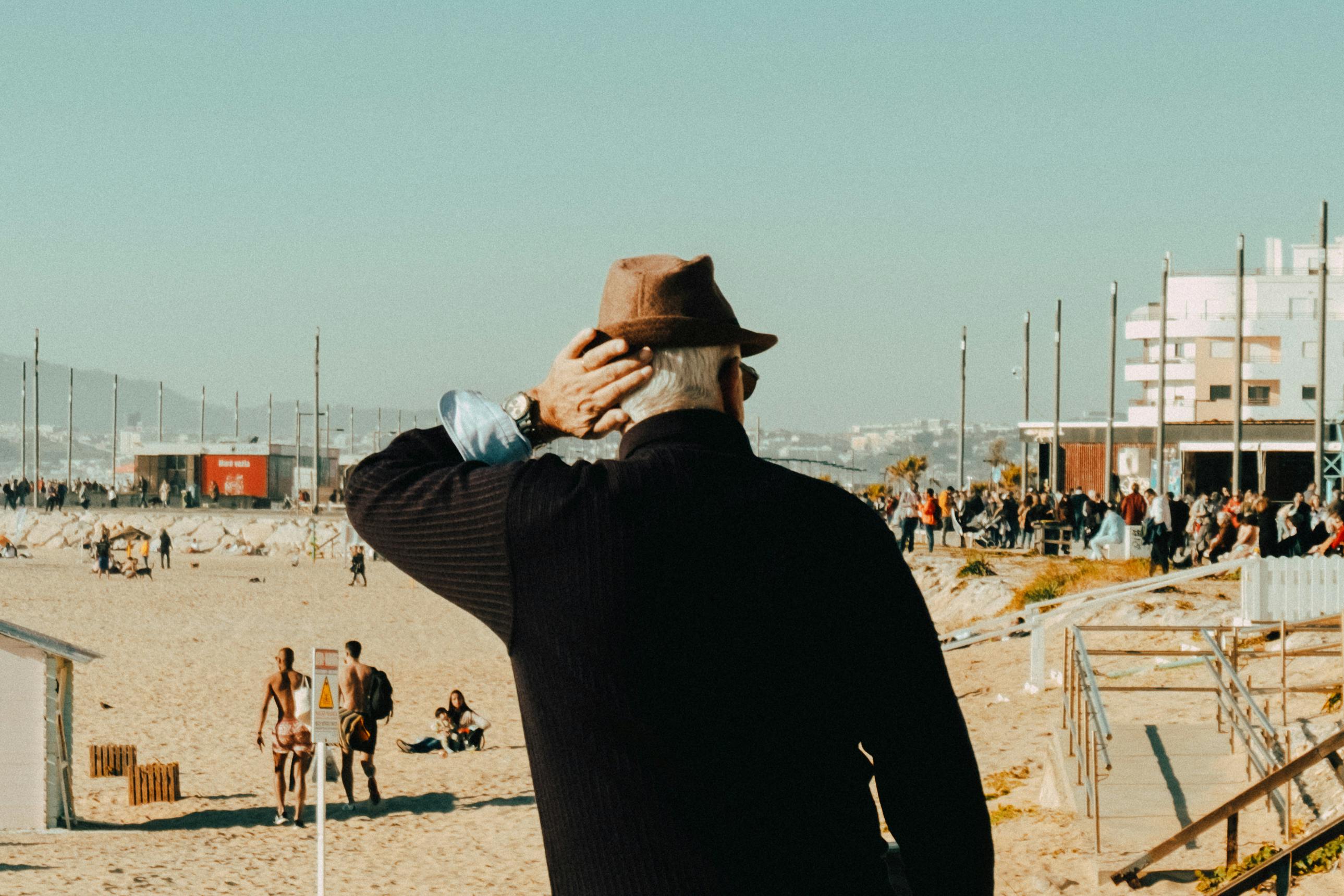 Senior man stands on Lisbon beach, enjoying the sunny day. Scenic view of the city with people relaxing nearby.