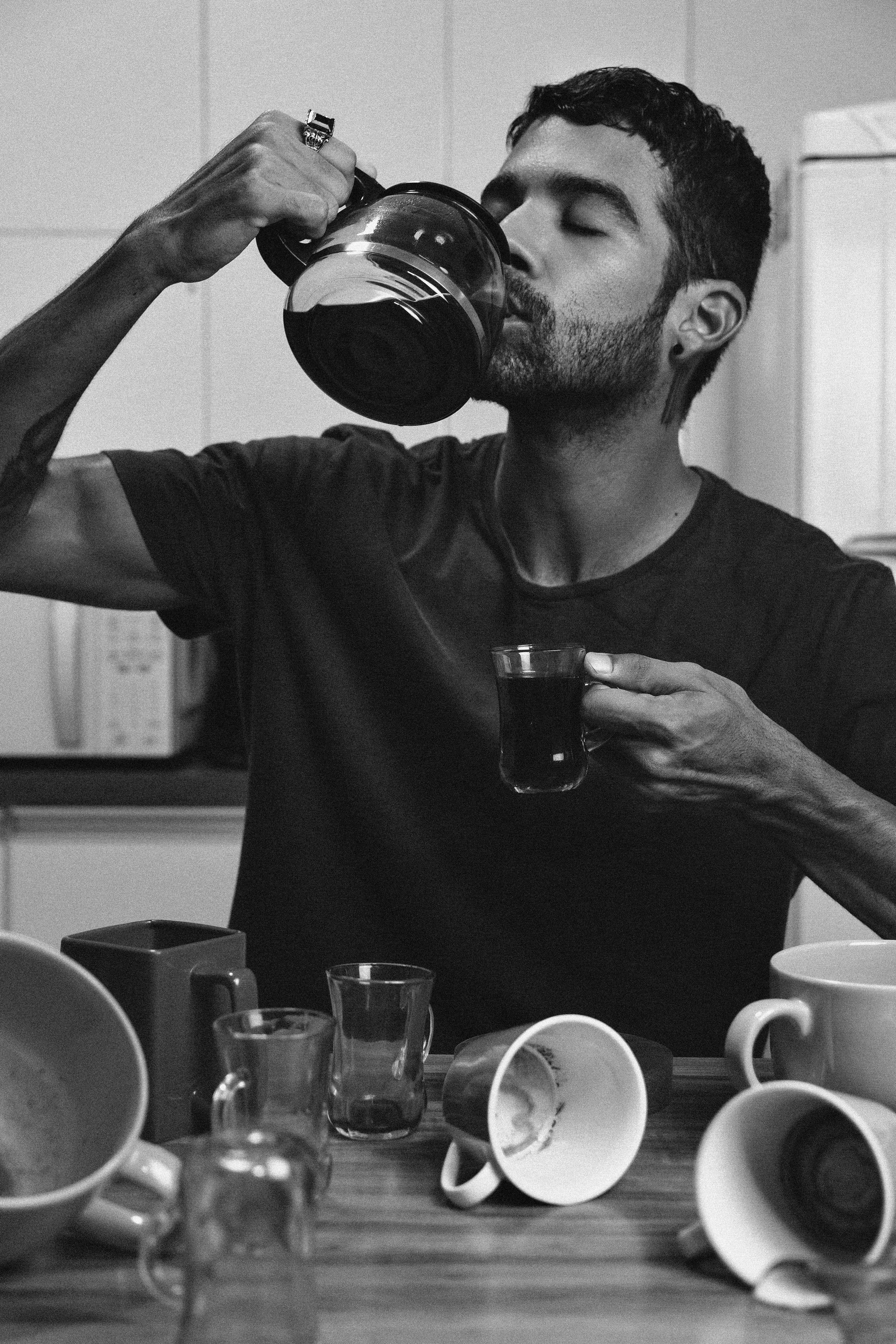 Black and white portrait of a man savoring coffee with an array of cups on a table, creating a vibrant yet minimalist aesthetic.