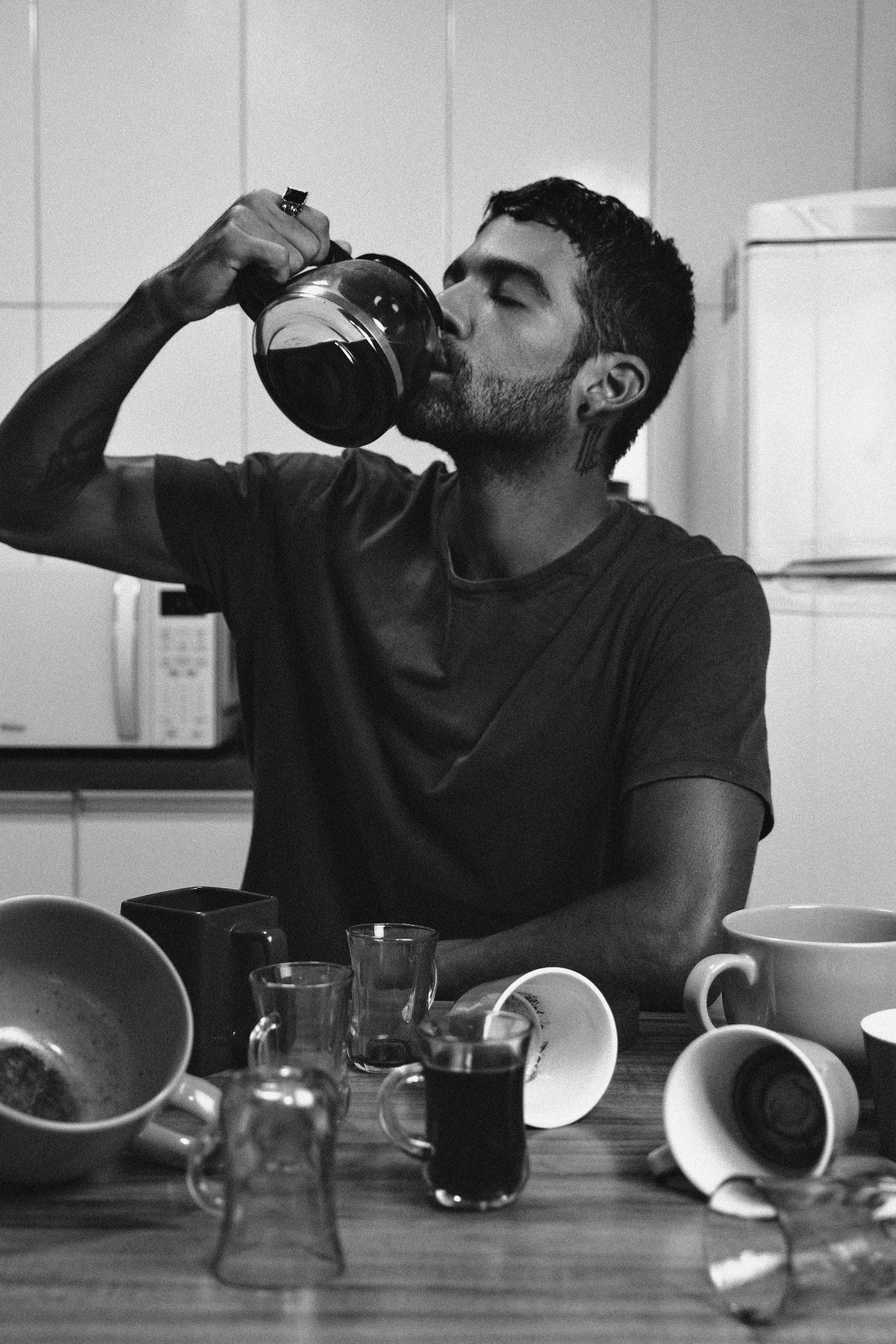 Black and white photo of a man drinking coffee straight from a pitcher at the kitchen table.