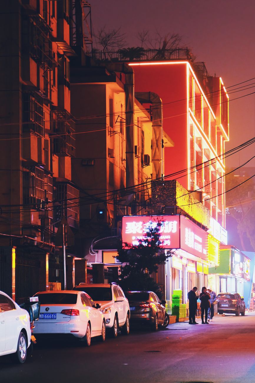 Illuminated city street at night with cars parked and vibrant neon signs creating a dynamic urban scene.