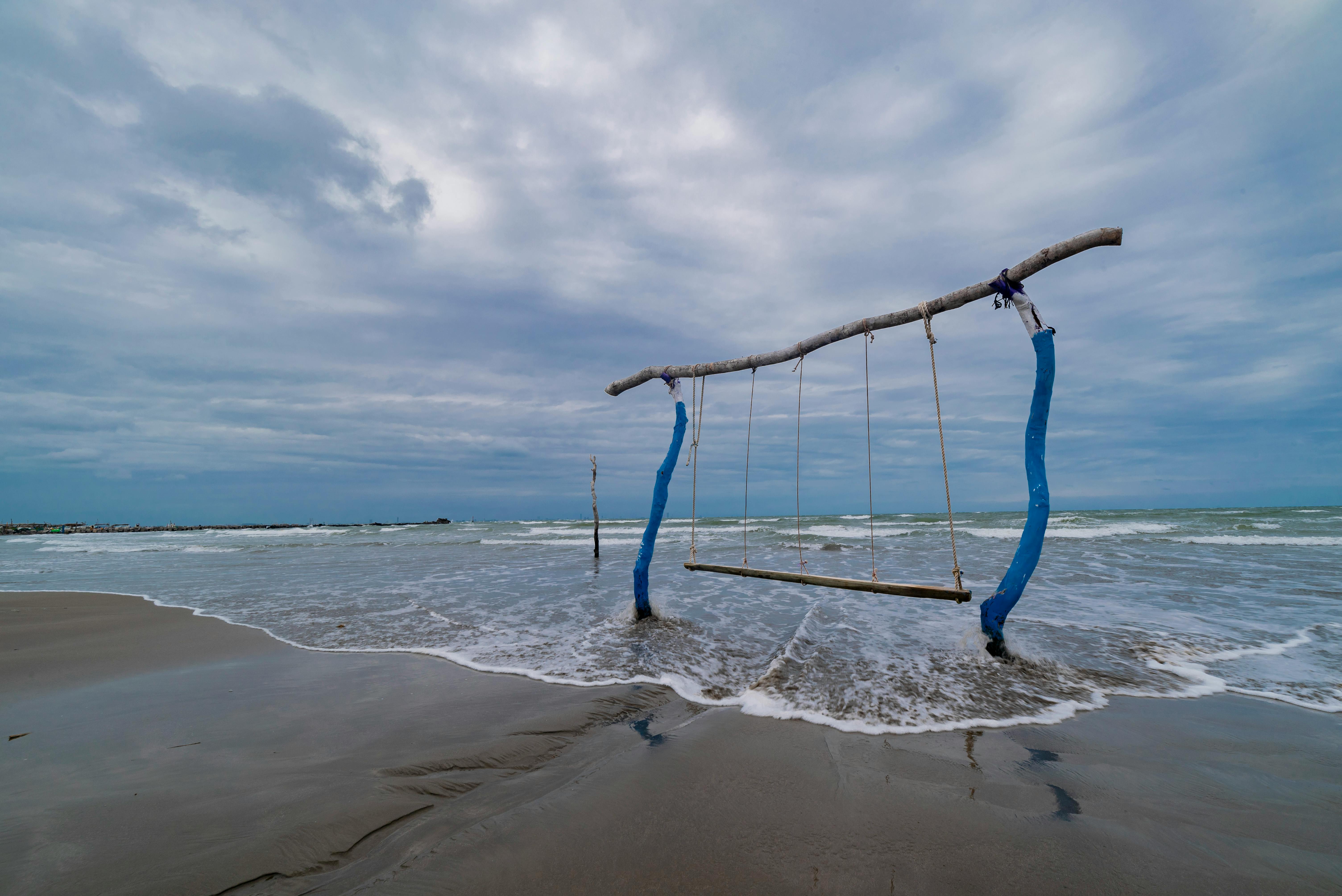 A rustic swing set over gentle waves on Boca del Río Beach, Veracruz, under a cloudy sky.