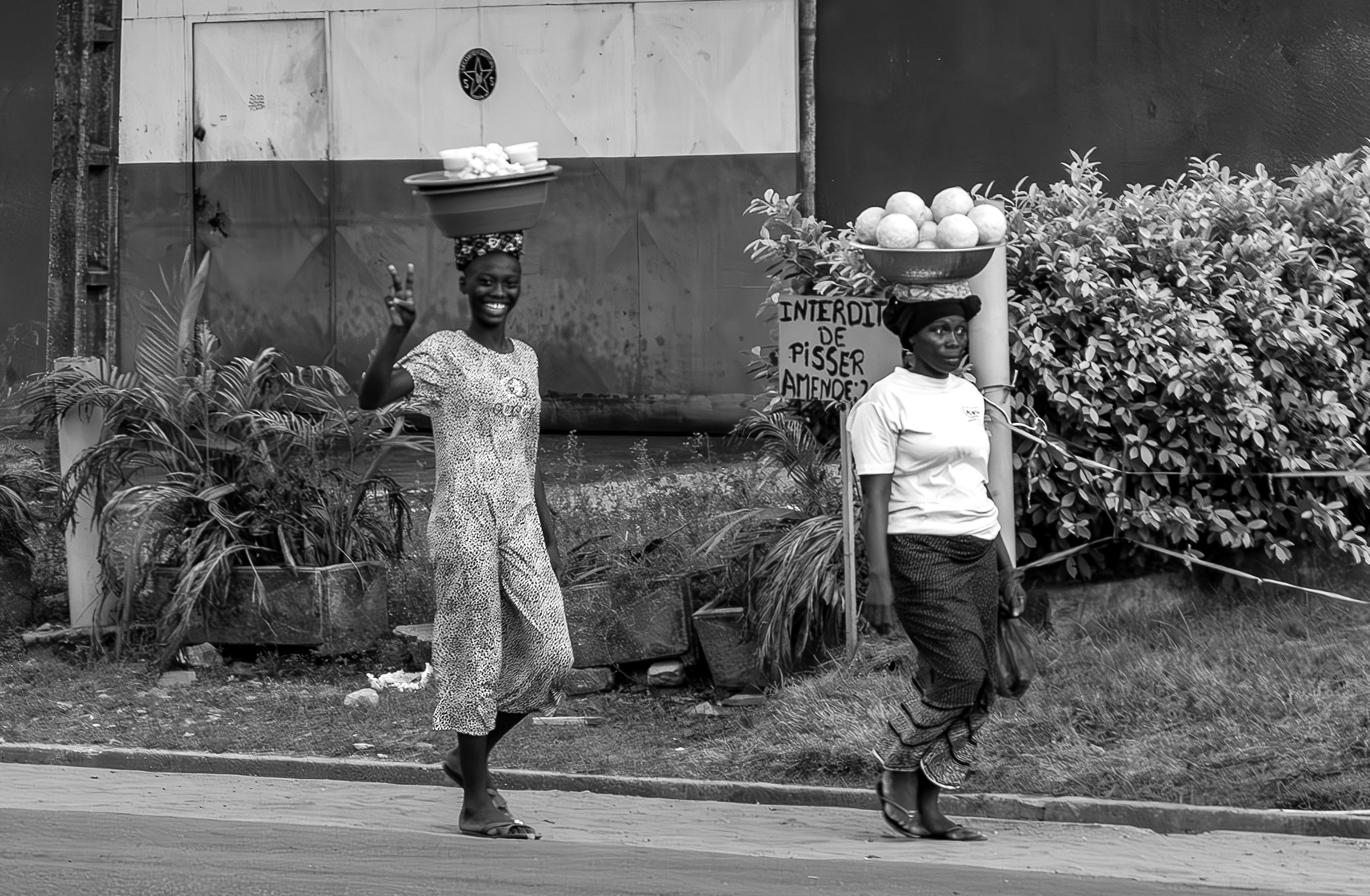 Two women carrying goods on their heads in a bustling urban street environment.