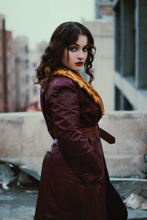 Free Fashionable woman in a red leather coat posing outdoors with a cityscape background. Stock Photo