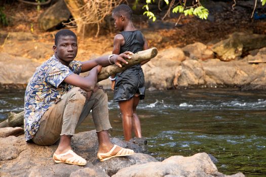 Two boys by a river, engaging with nature, sitting on rocks and holding a log.