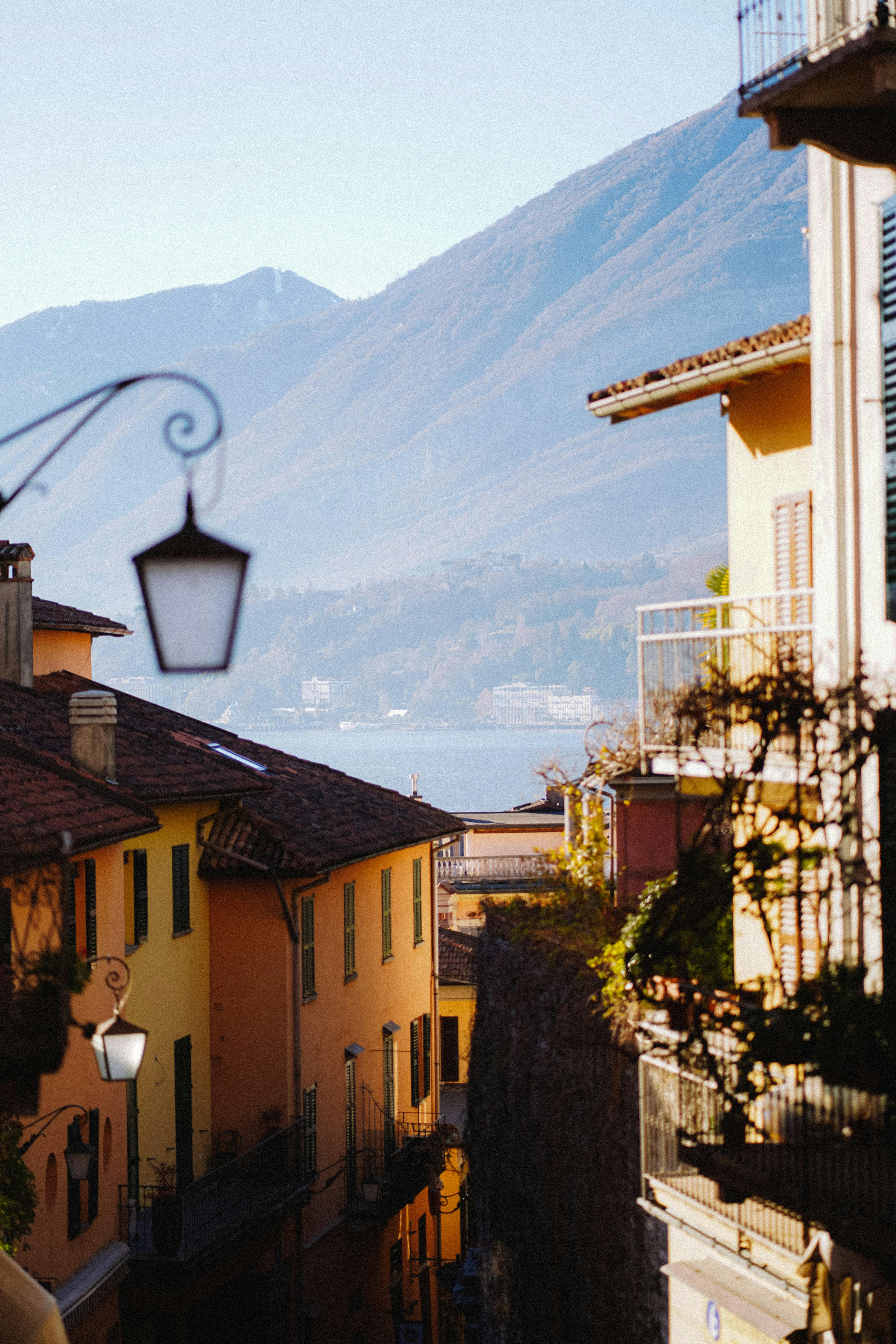 Sunlit view of an Italian town street with mountains and lake in the background, creating a picturesque scene.