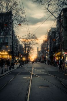 Bustling urban street with tram tracks at sunset, vibrant city life and architecture.