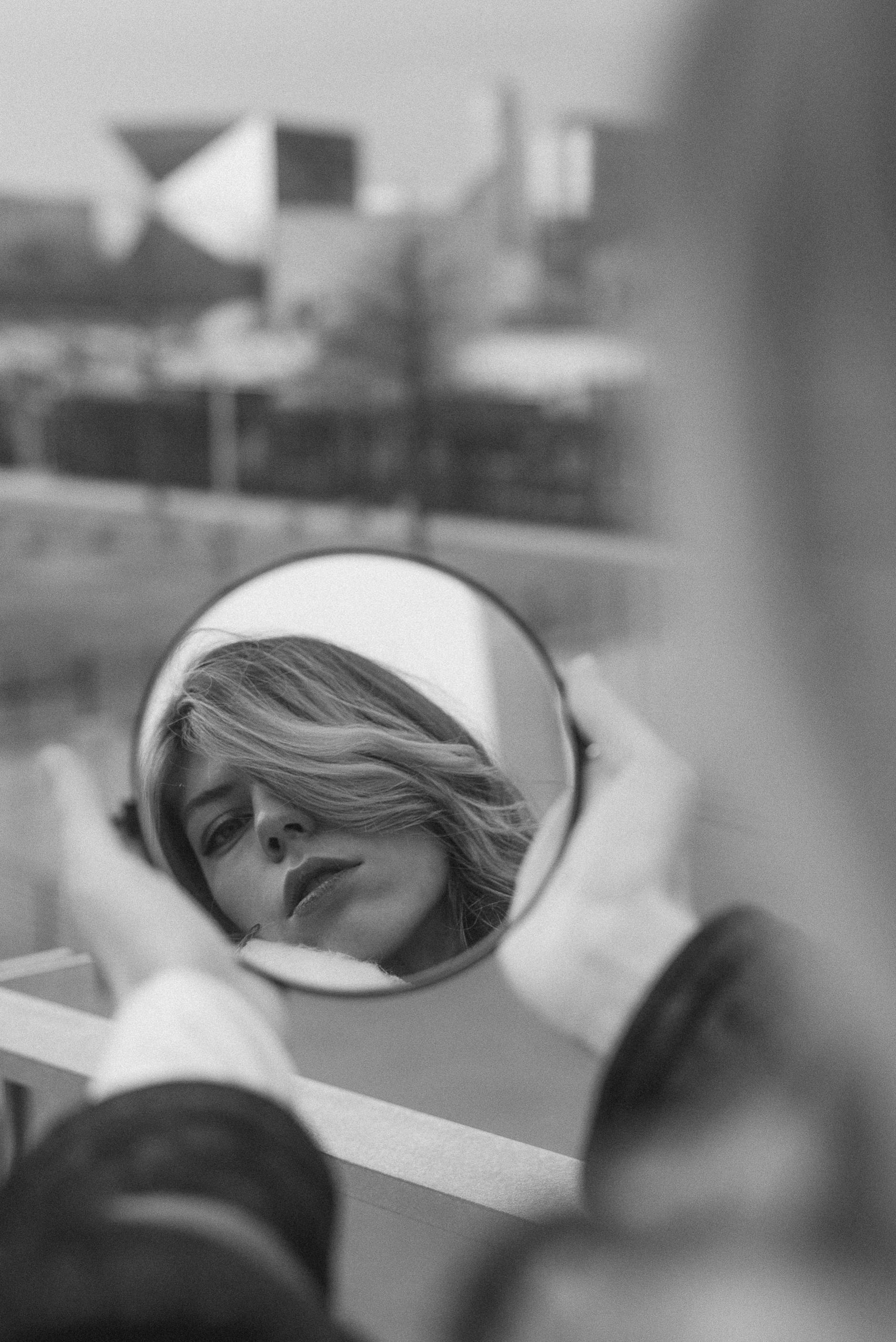 Artistic black and white portrait of a woman holding a mirror reflecting her face in an urban setting.