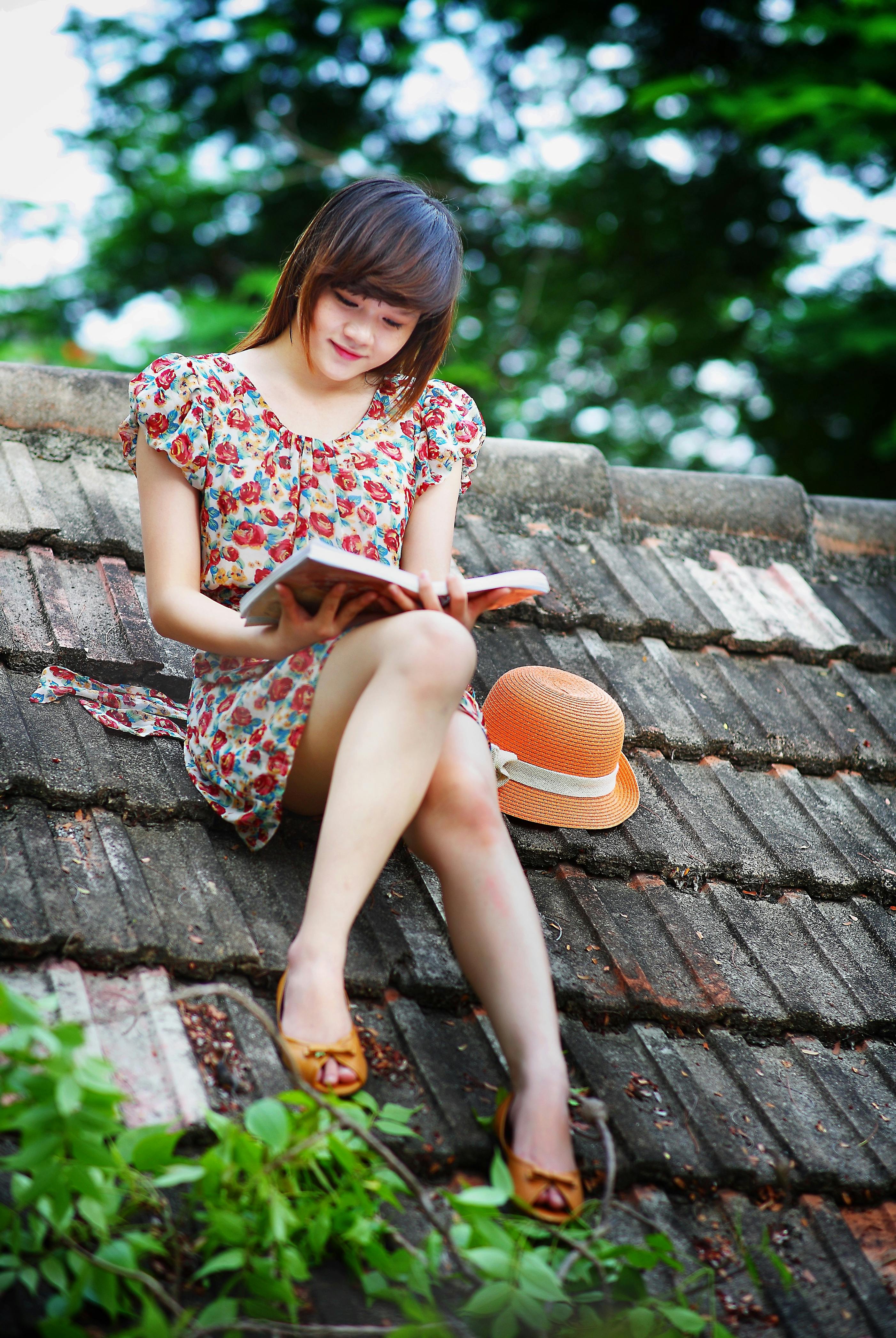 Woman Wearing White, Green, and Red Floral Cap-sleeved Dress Reading a Book on Black Roof Near Green Trees