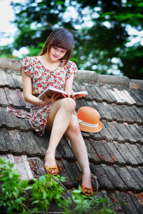 녹색 나무 근처에 검은 지붕에 책을 읽고 흰색, 녹색 및 빨간색 꽃 모자 소매 드레스를 입고 여자