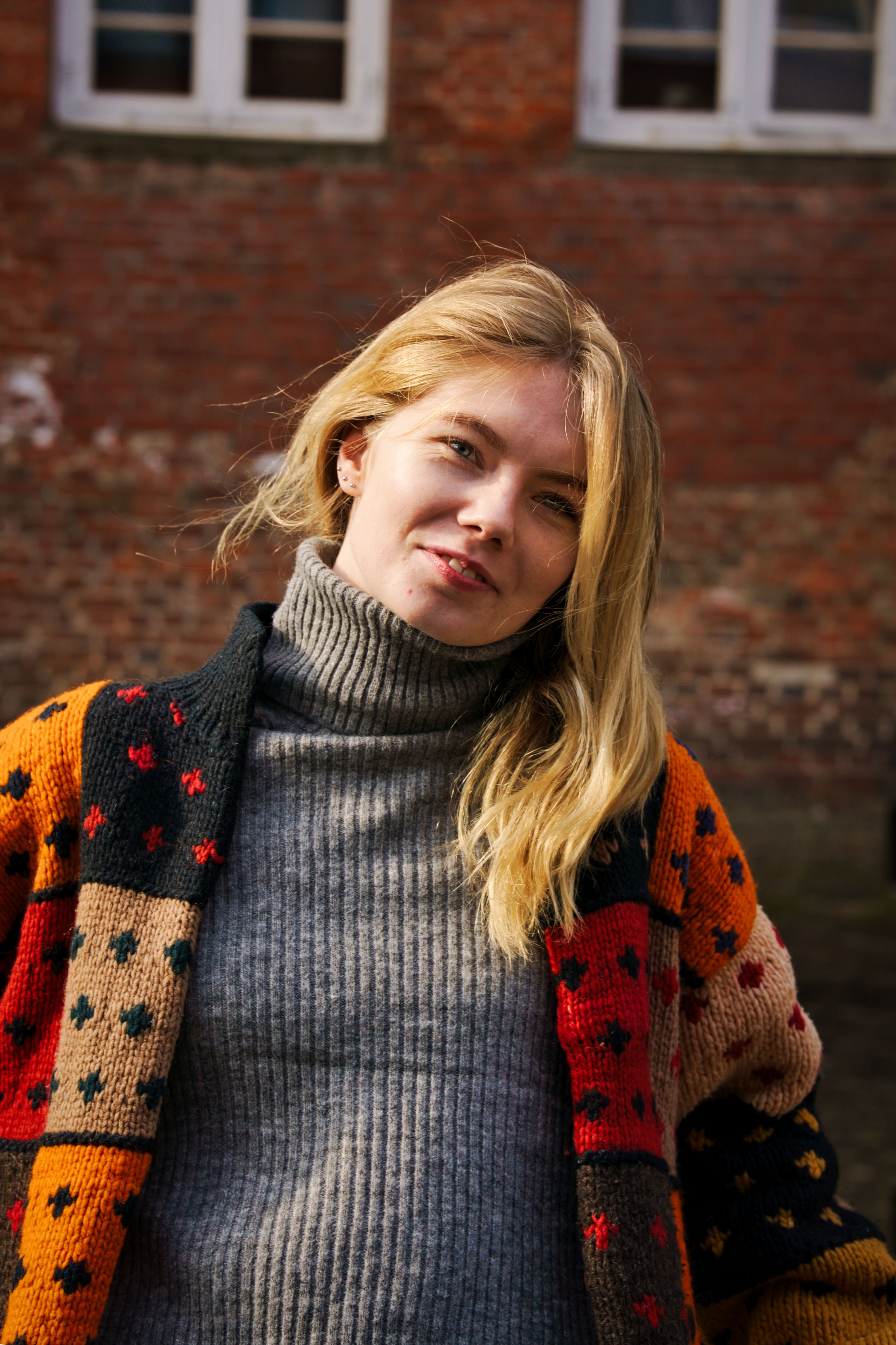 A young blonde woman in a colorful sweater poses outdoors against a brick wall.