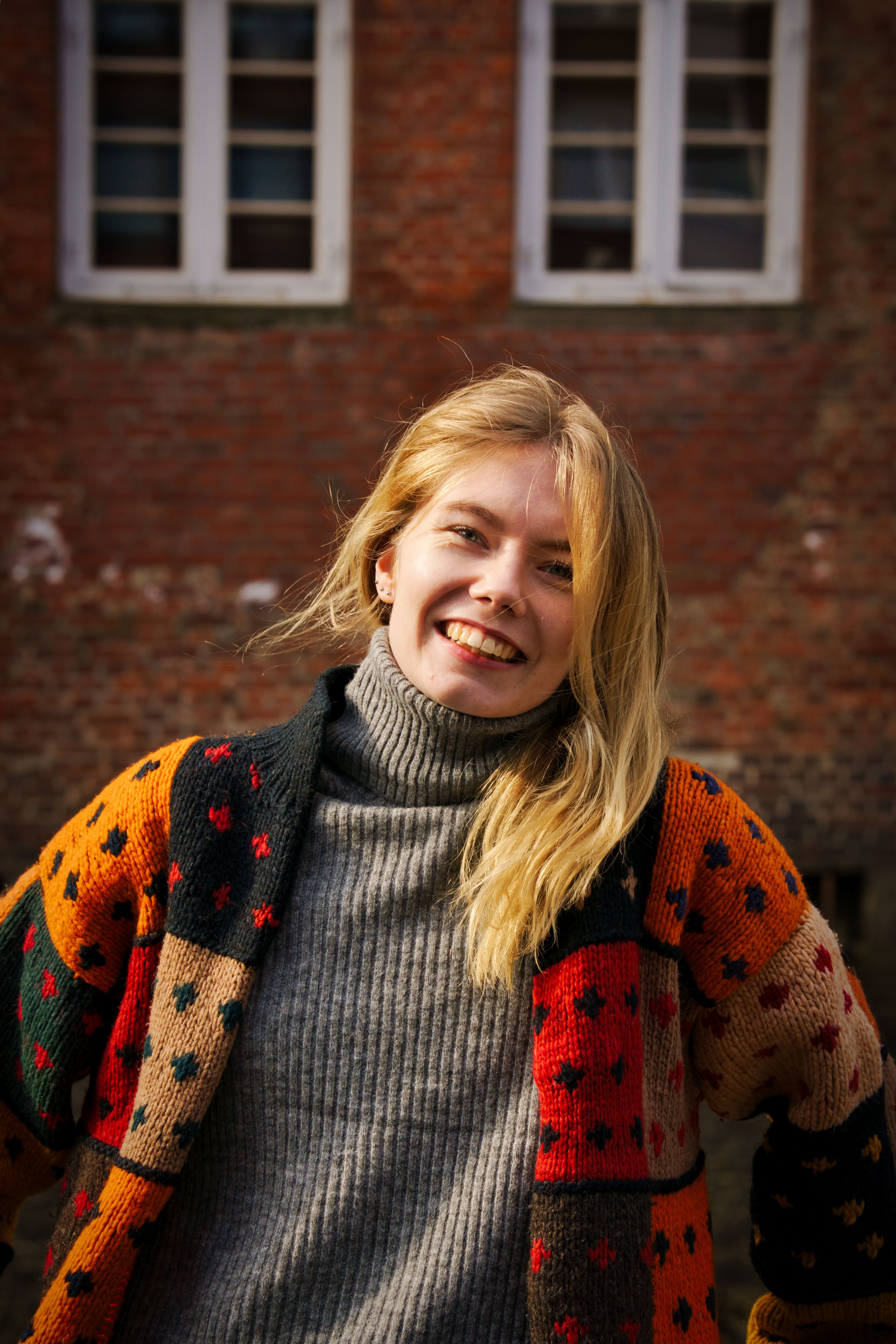 Smiling woman with blonde hair wearing a cozy multicolored sweater against a brick wall background.