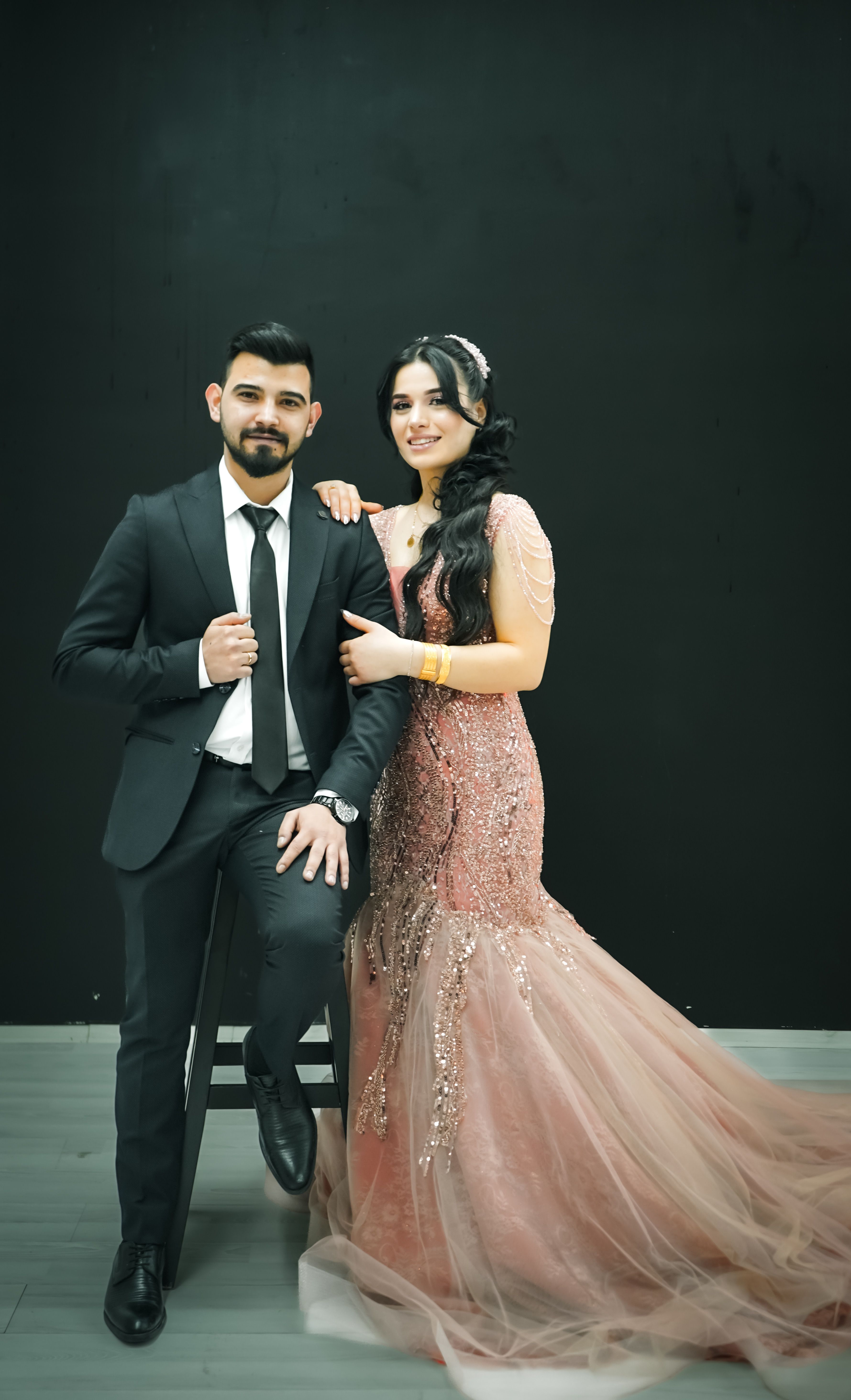 A couple elegantly dressed in a black suit and pink dress, posing against a black background.