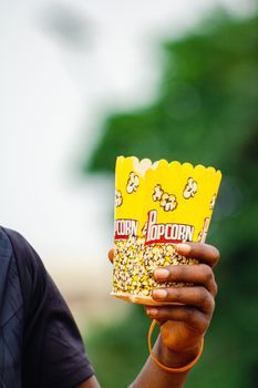 Close-up of a hand holding a yellow popcorn box outdoors, vibrant colors.