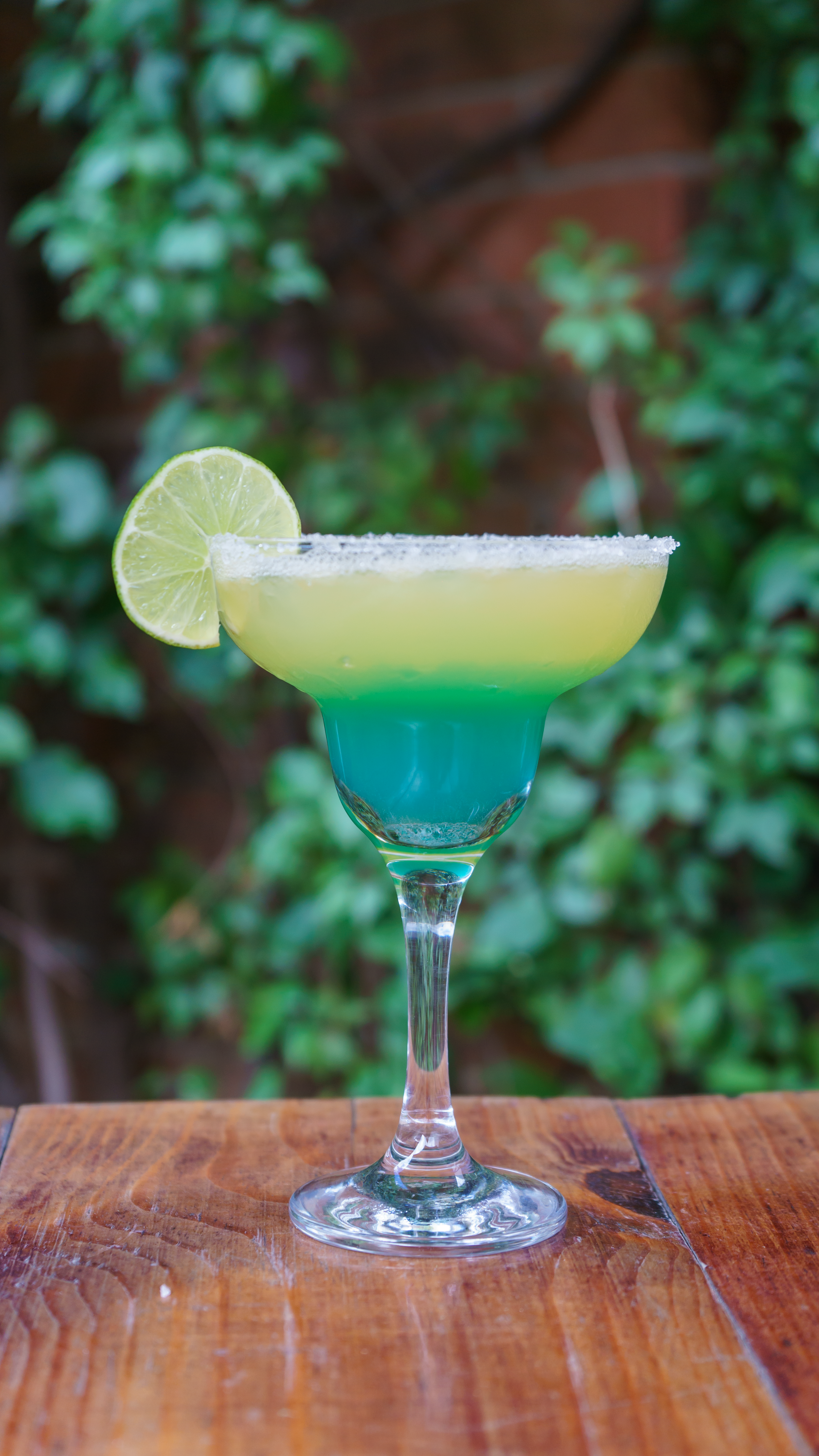 Refreshing margarita cocktail with lime on a wooden table against a lush background.