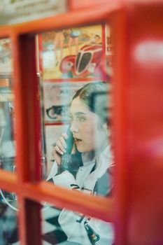 A young woman talking on a pay phone inside a vintage red telephone booth.