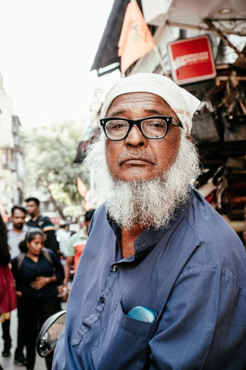 Free Portrait of an elderly man with a white beard and glasses in a busy Mumbai street market. Stock Photo