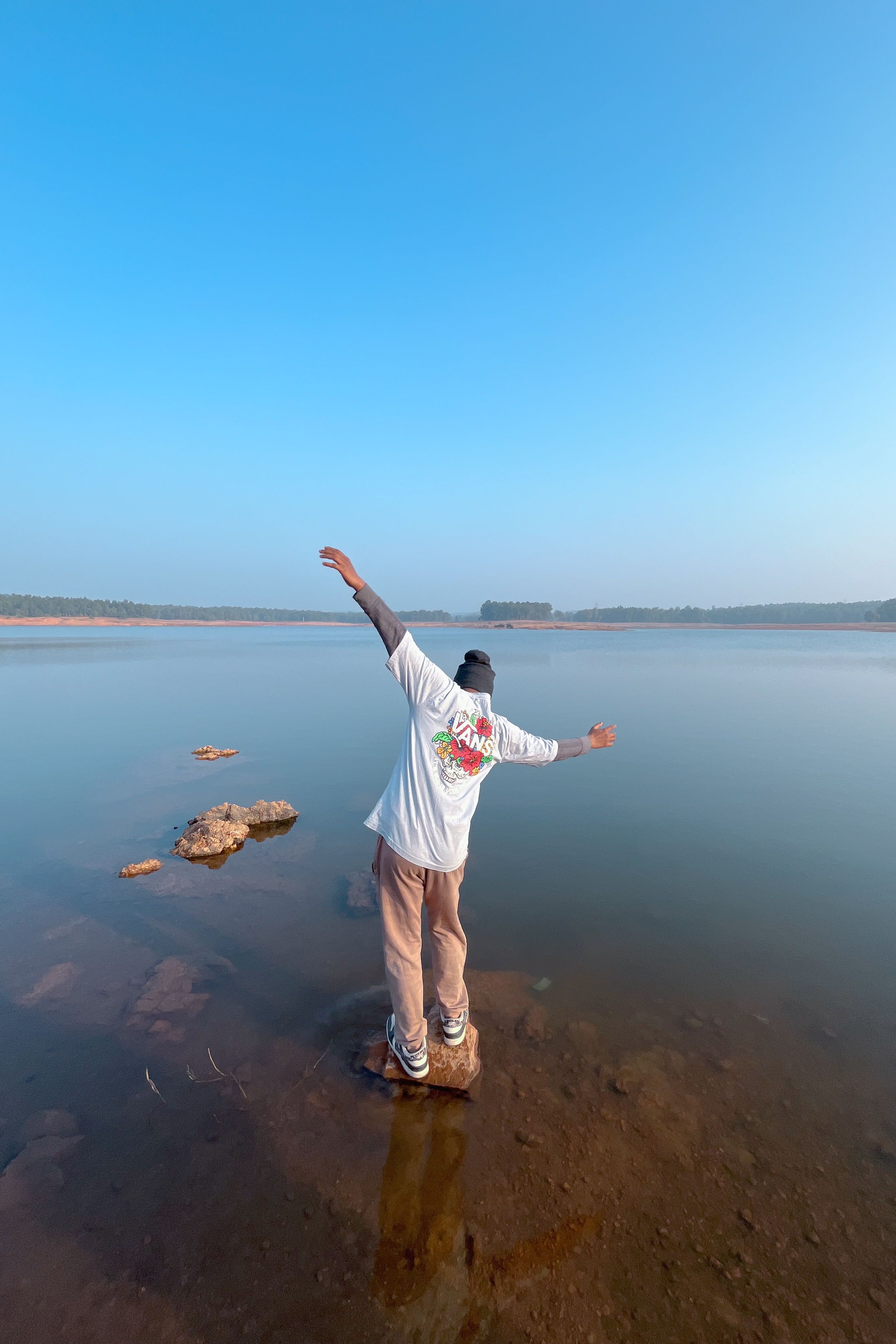 A man stands on rocks, arms spread, overlooking a calm lake under a clear blue sky in Hazaribagh, India.