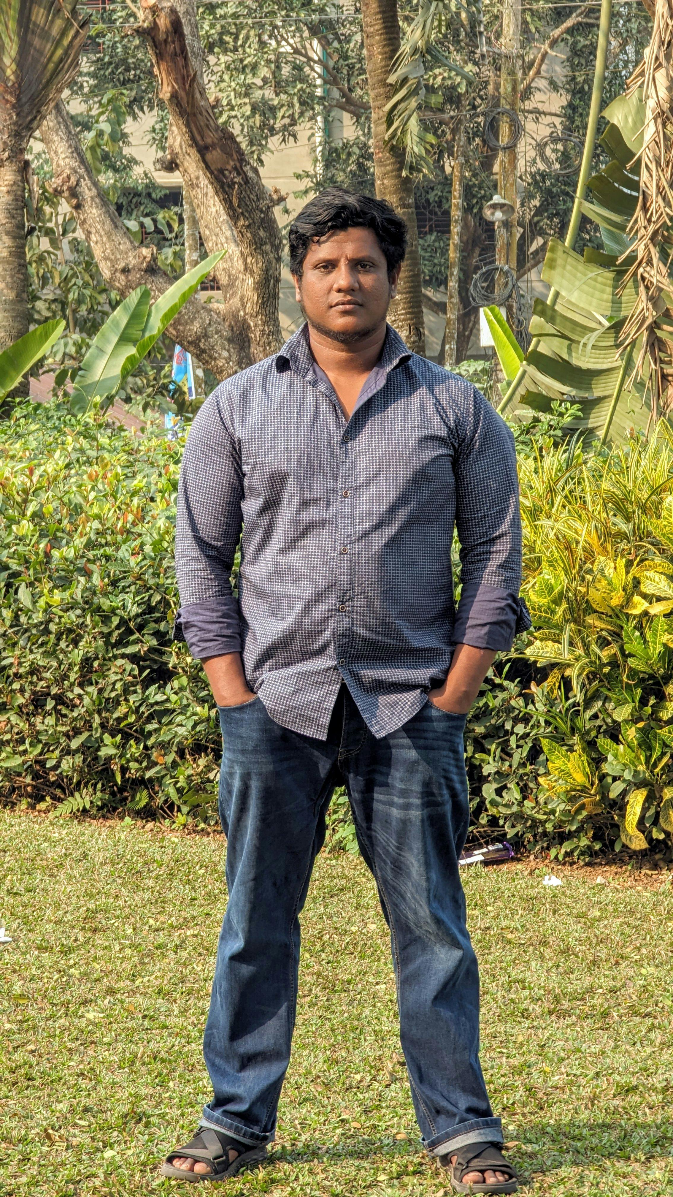 A man standing casually in a park in Chittagong, Bangladesh, surrounded by greenery.