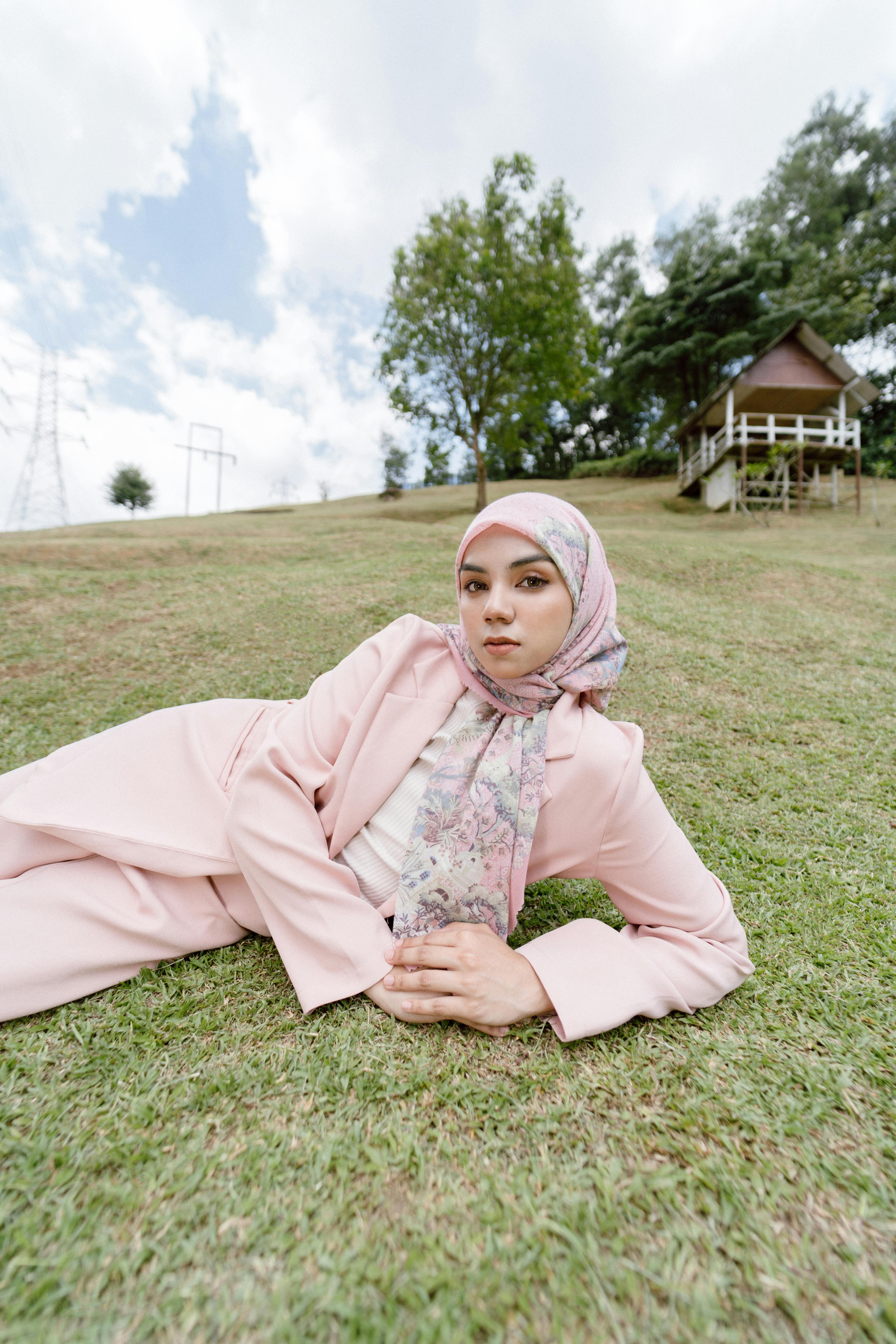 A fashionable woman in a pink suit and headscarf rests on a grassy meadow in Kuala Lumpur.
