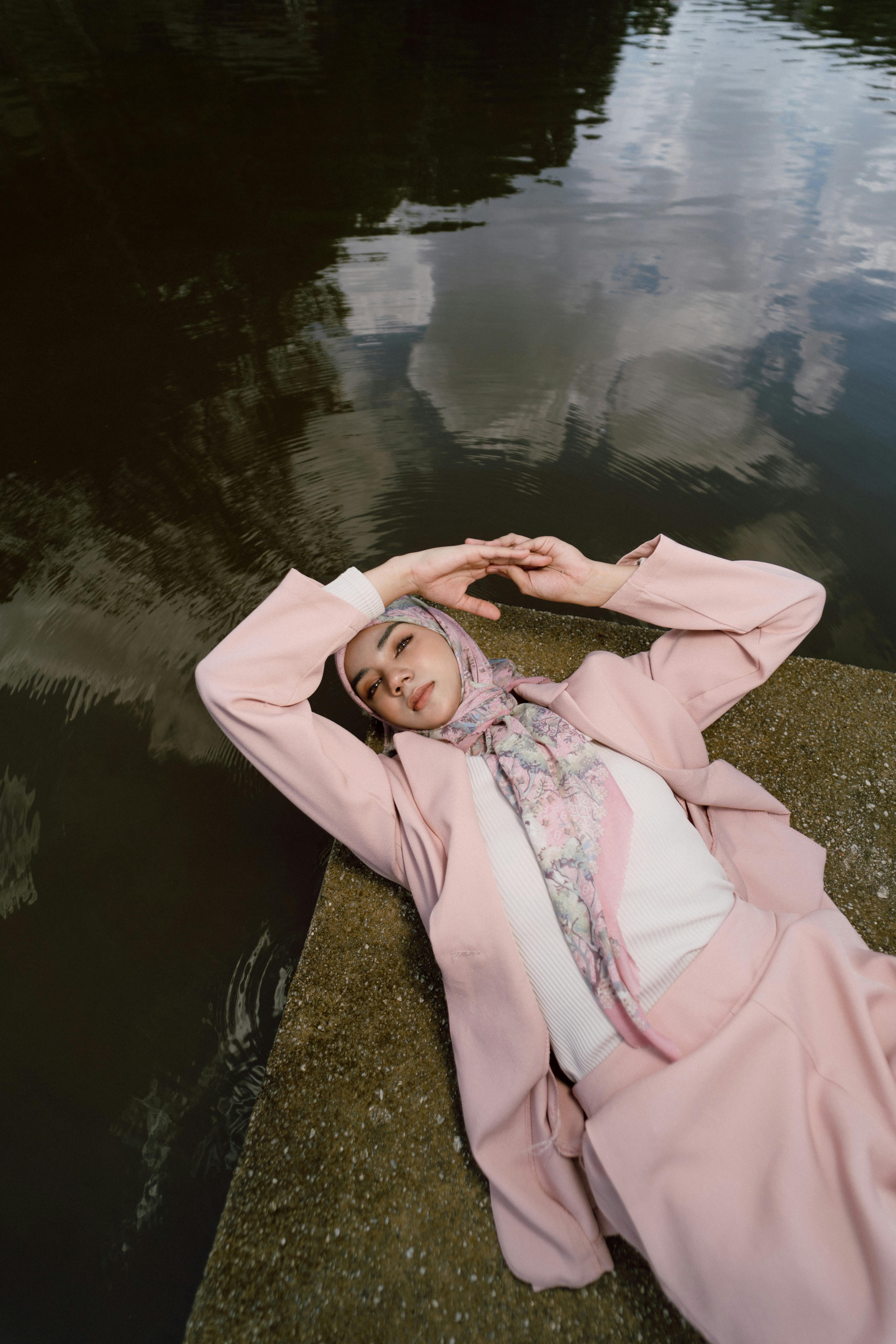 Elegant woman in a pink suit and hijab lying by a lakeside, captured from above.