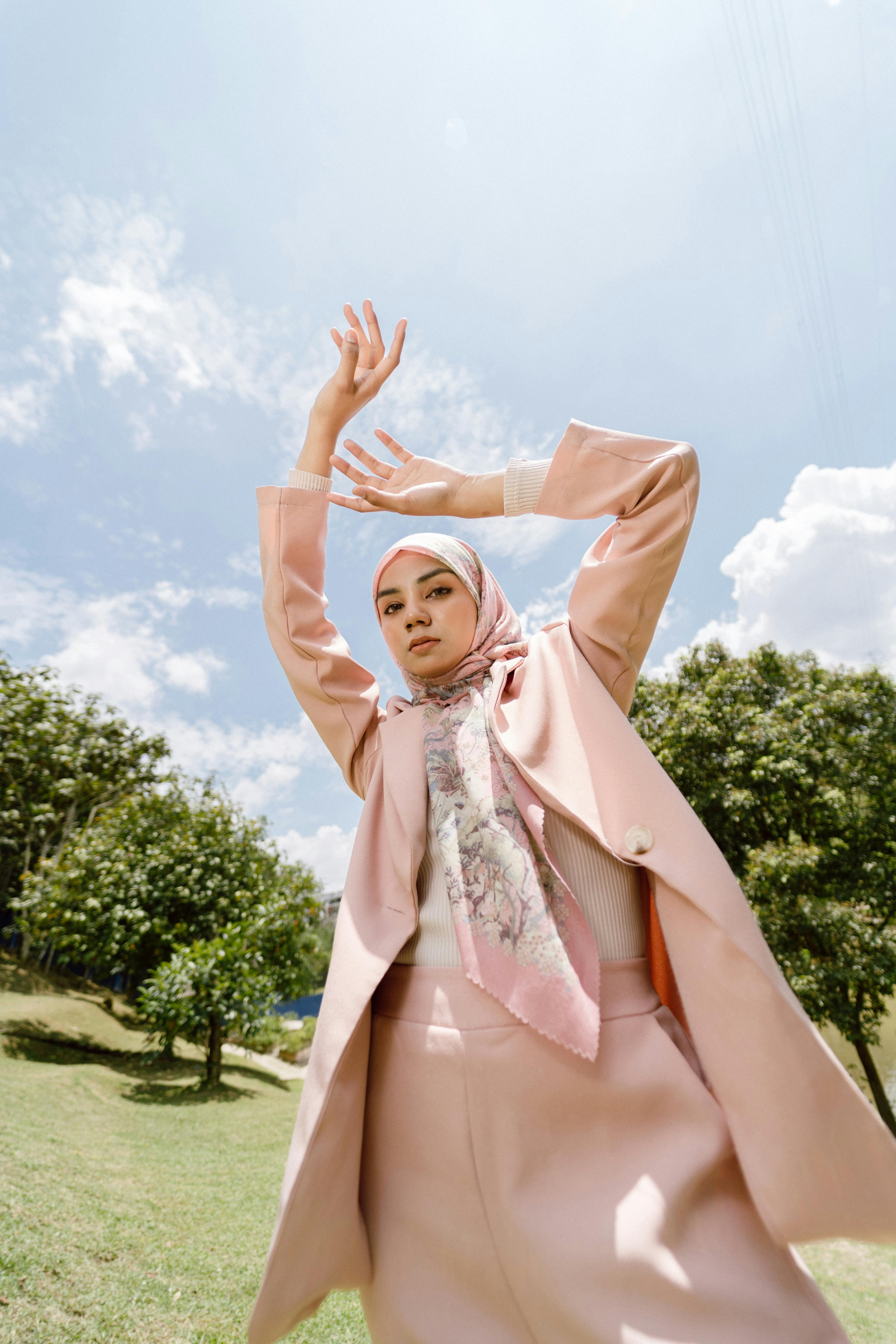 Stylish woman in a pink suit and hijab posing elegantly outdoors against a blue sky.