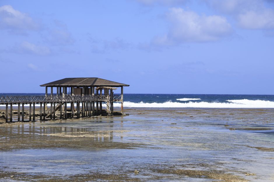 A serene view of a thatched-roof pier over calm ocean waters with waves and clear blue sky.