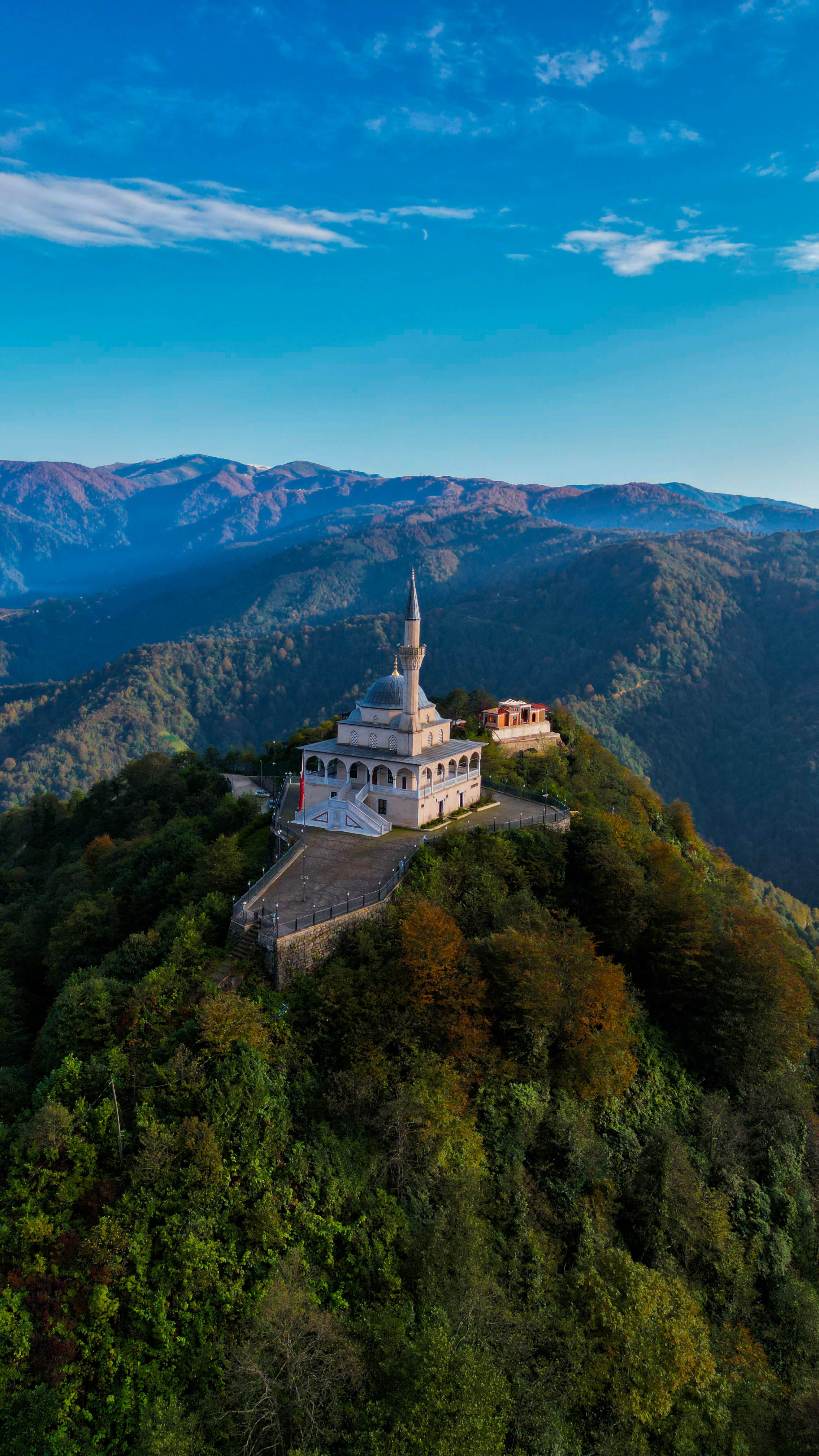 An aerial photograph capturing a stunning hilltop mosque surrounded by lush green forests against a mountain backdrop at sunrise.