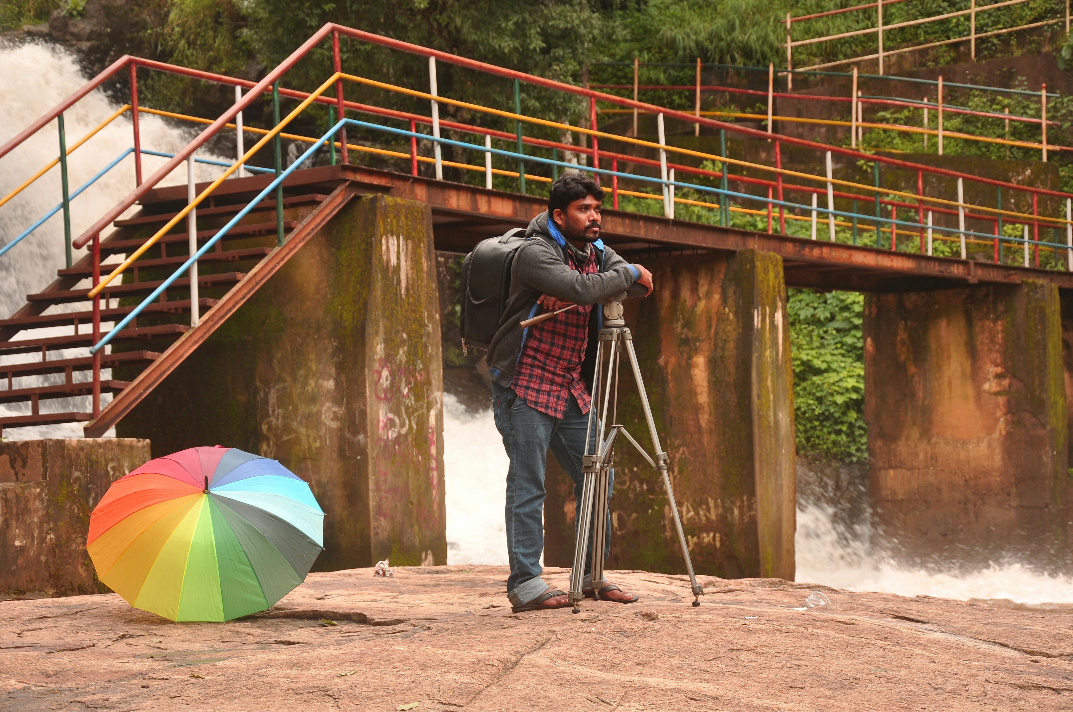 Man with tripod and umbrella near a vibrant bridge at a waterfall.