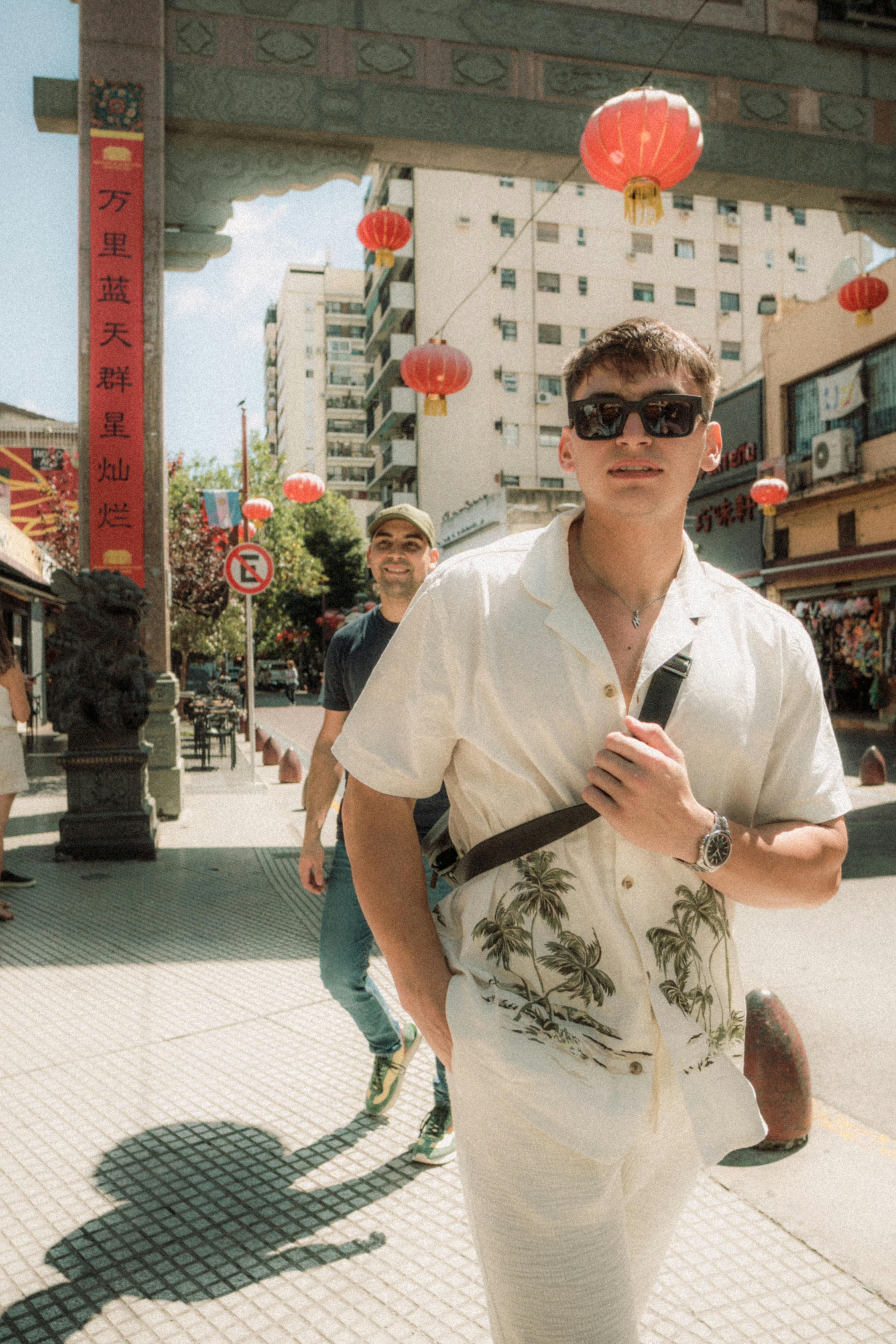 Two men walking under red lanterns in an urban Chinatown setting, wearing casual summer attire.