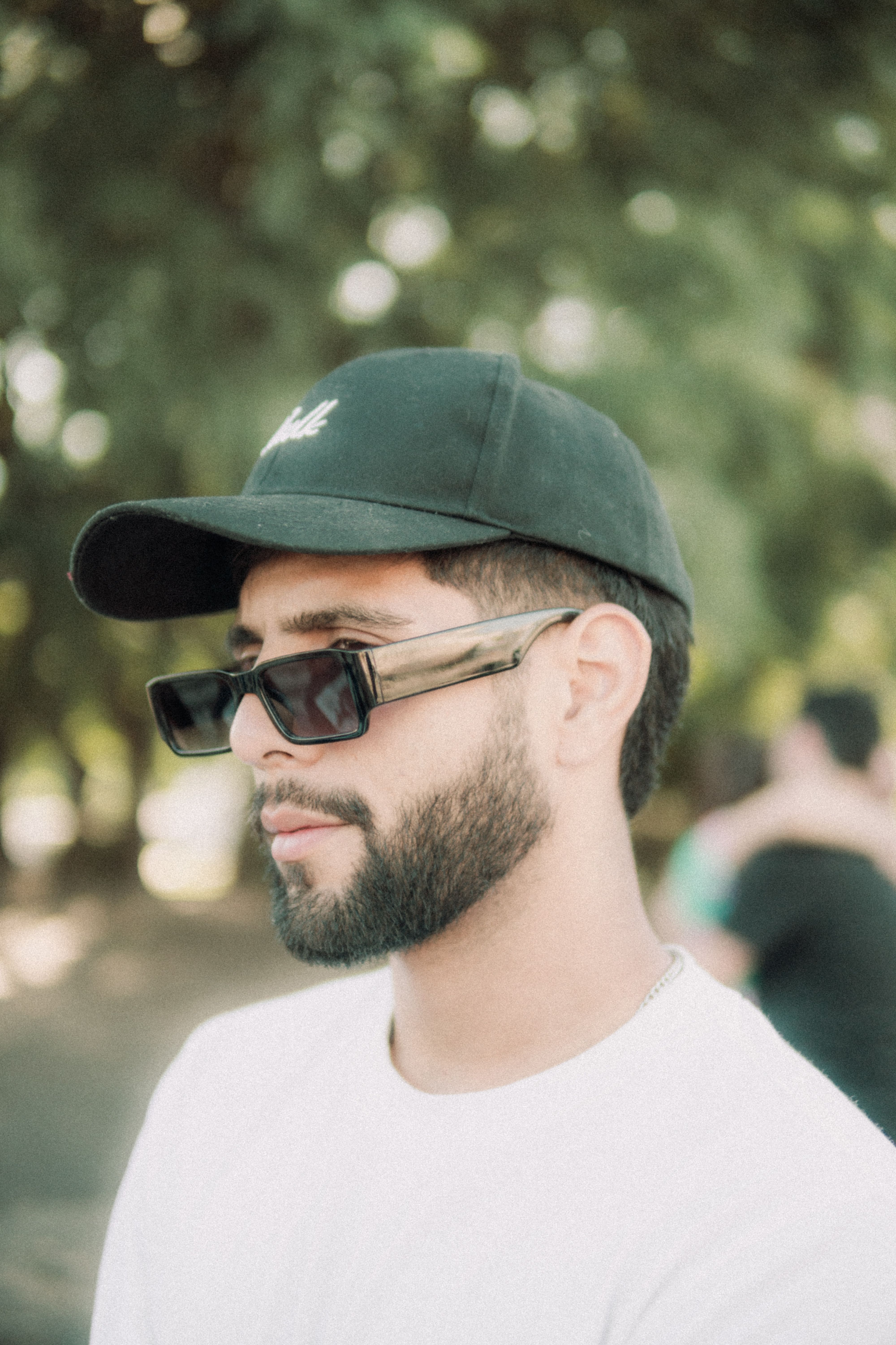 Portrait of a bearded man wearing a black cap and sunglasses in a natural outdoor setting.