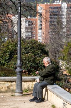 Senior man sitting in a city park, checking his watch under a lamppost.