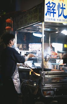 A street food vendor prepares food at a bustling night market, illuminated by bright lights.