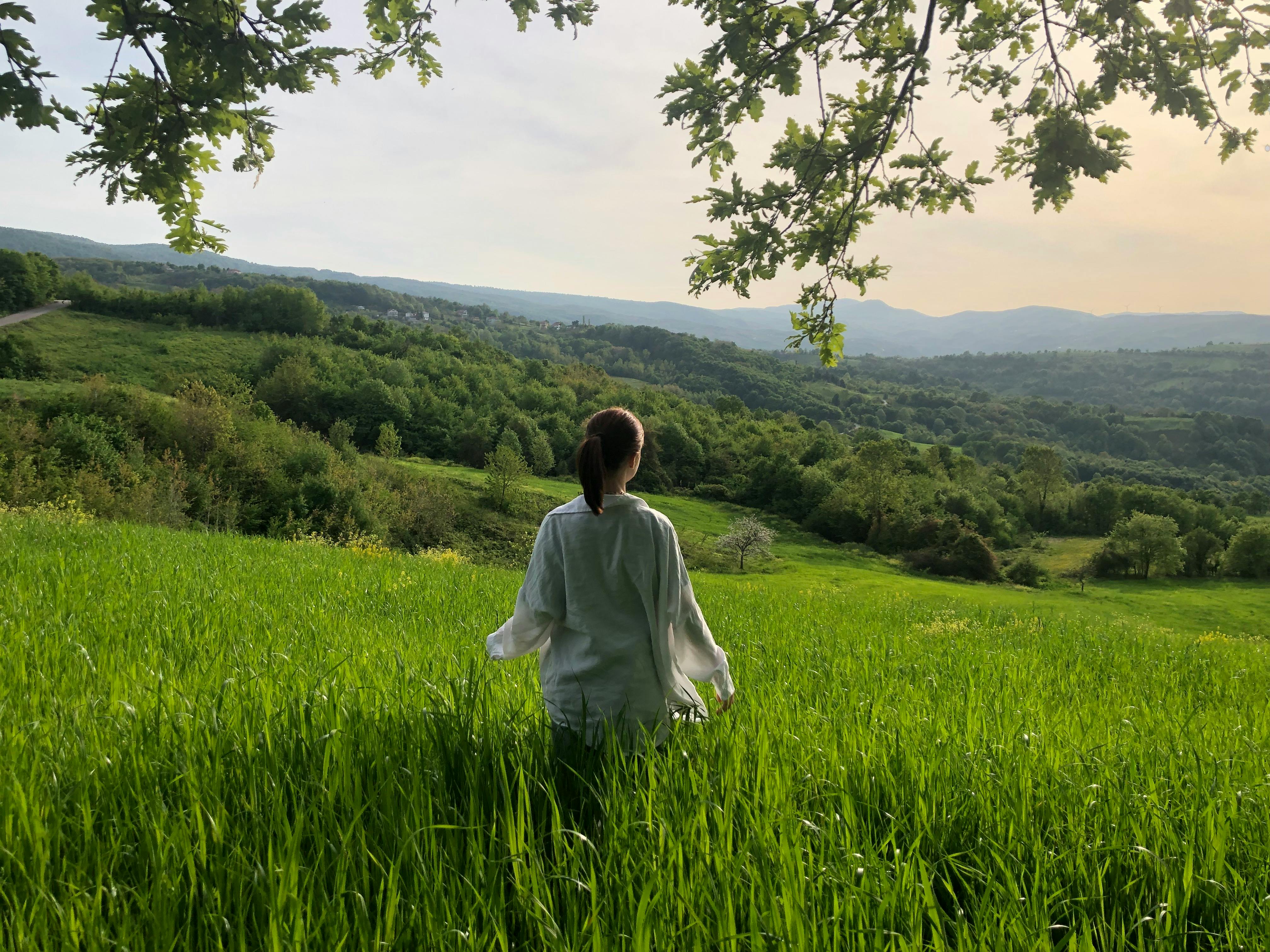 A woman stands in a lush green field, surrounded by rolling hills and forests, enjoying nature's serenity.