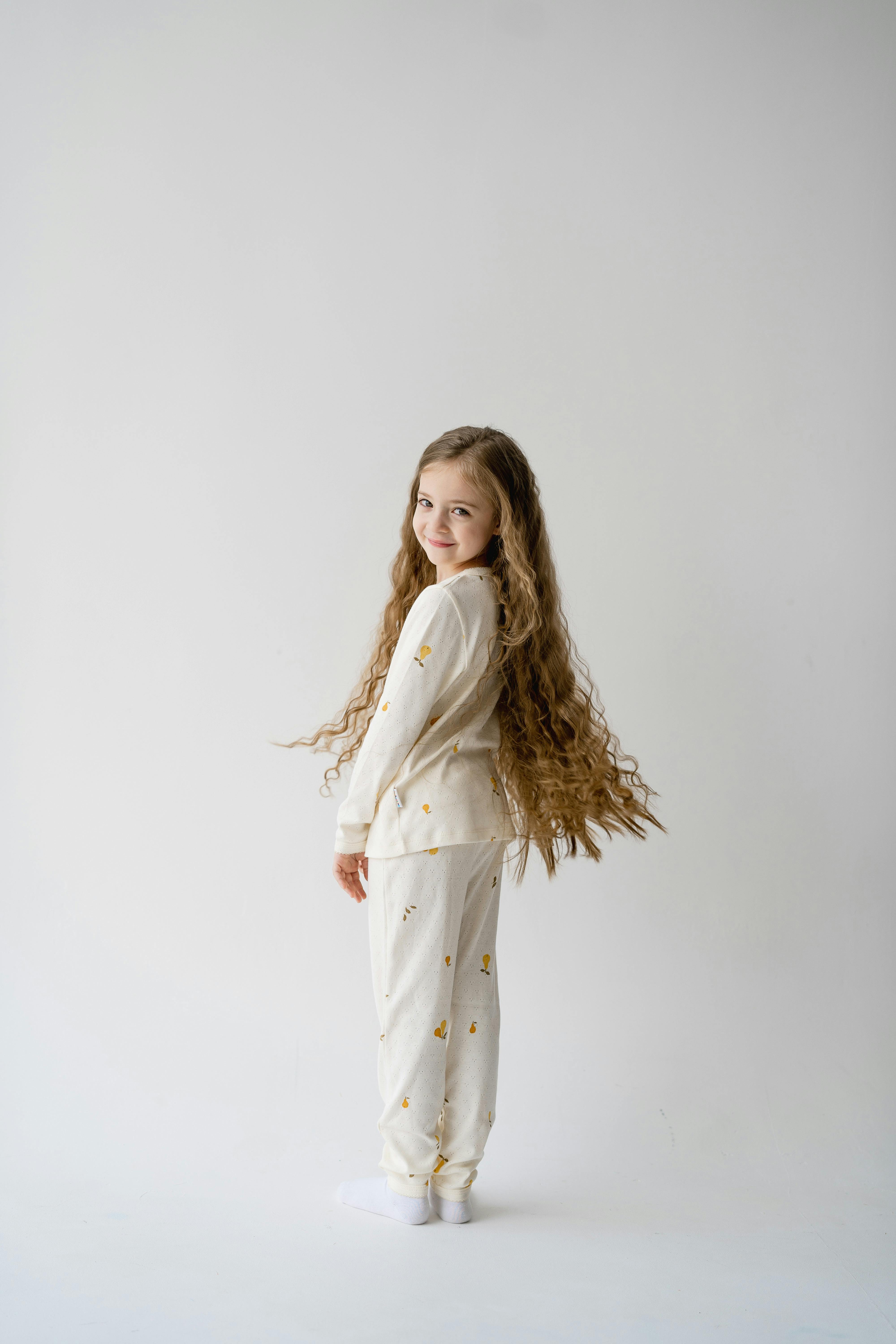 A young girl with long hair wearing pajamas poses against a white background, smiling over her shoulder.